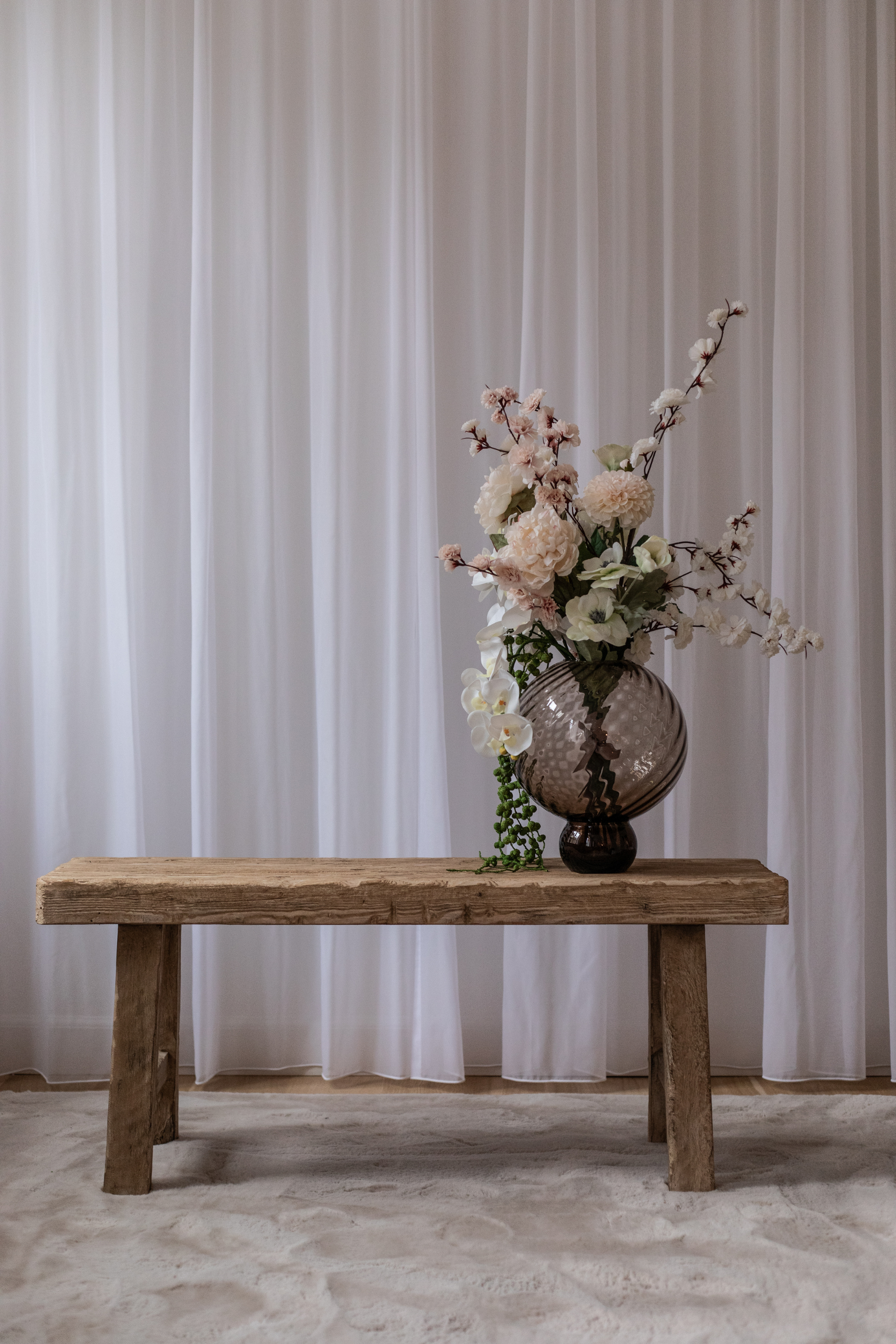 En rustik træbænk står foran hvide gardiner med en stor rund glasvase ovenpå. Vasen er fyldt med lyse blomster og grønne grene, som hænger ned over bænken.