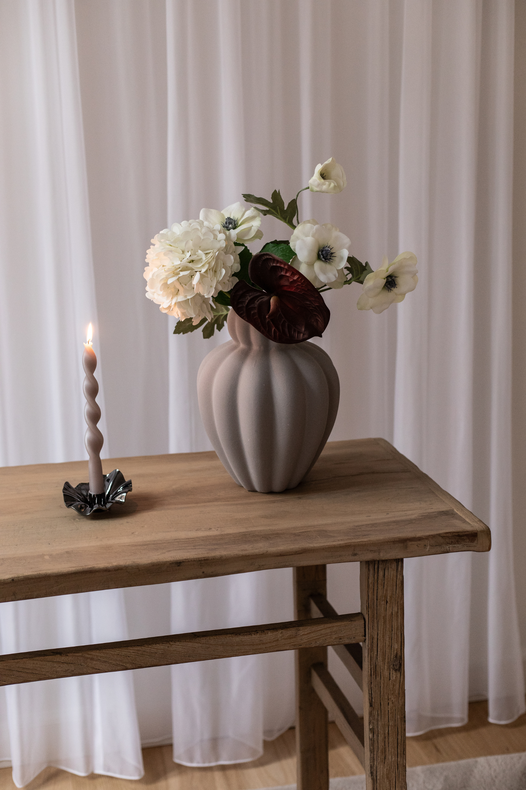 En grå, riflet vase med hvide og mørkerøde blomster står på et rustikt træbord. Ved siden af står en snoet tændt lysestage med mørk holder foran hvide gardiner.