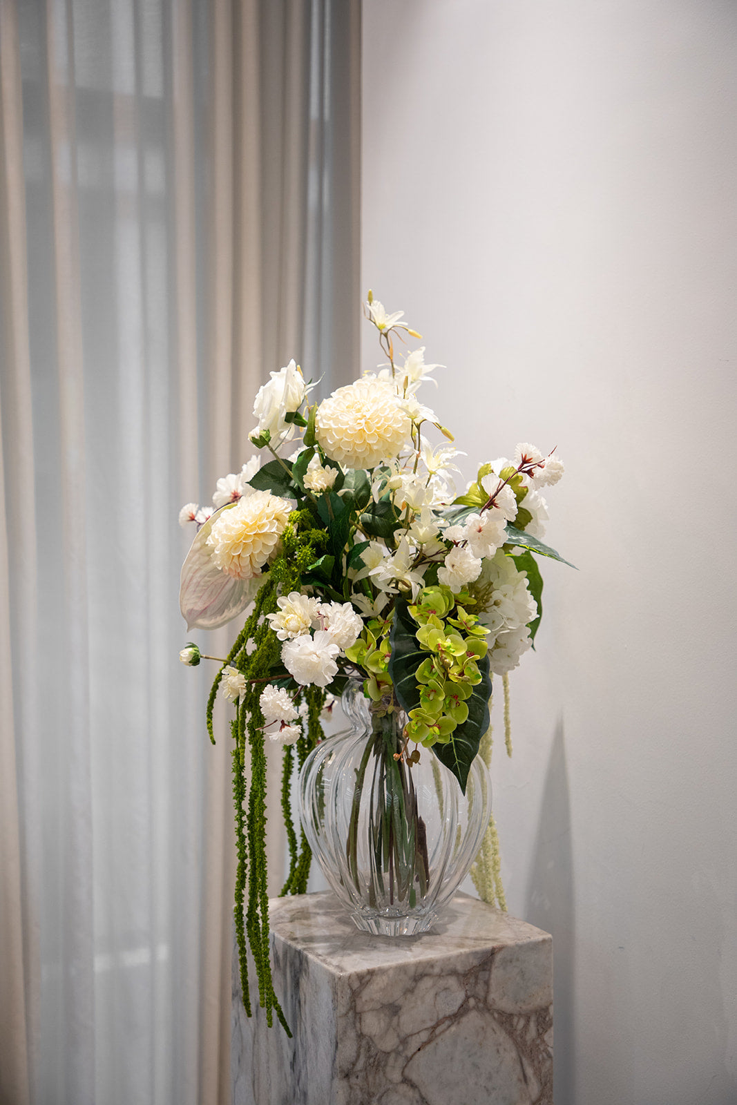 Stor buket af kunstige blomster i hvide og grønne nuancer. Placeret i en gennemsigtig glasvase, der står oven på en marmorpedestal.
