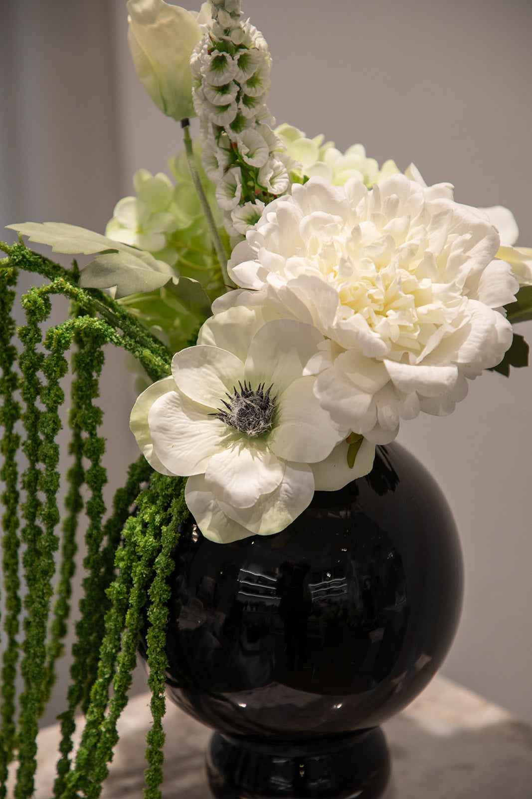 Nærbillede af buket af kunstige blomster i hvide og grønne nuancer. Placeret i en blå glasvase, der står oven på en marmor pedestal.