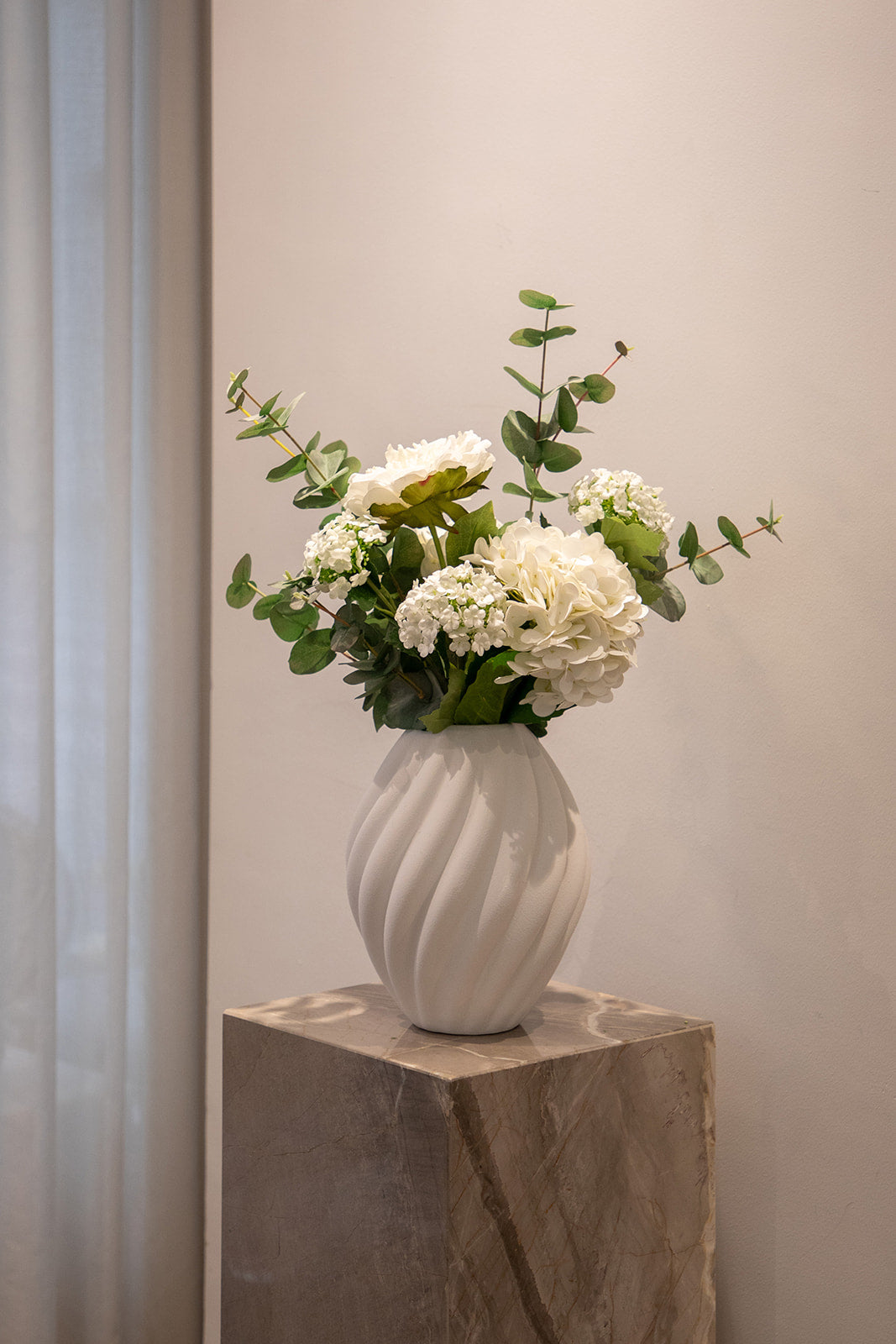 Basis buket af kunstige blomster i hvide og grønne nuancer, placeret i en hvid keramik vase. Vasen står på en marmor piedestal.
