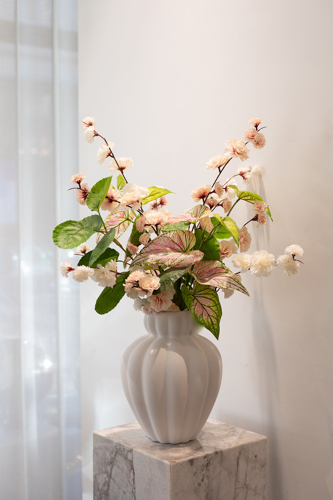 Kunstig buket i sarte rosa, hvide og grønne nuancer. Placeret i en hvid keramik vase, der står på en marmor pedestal.