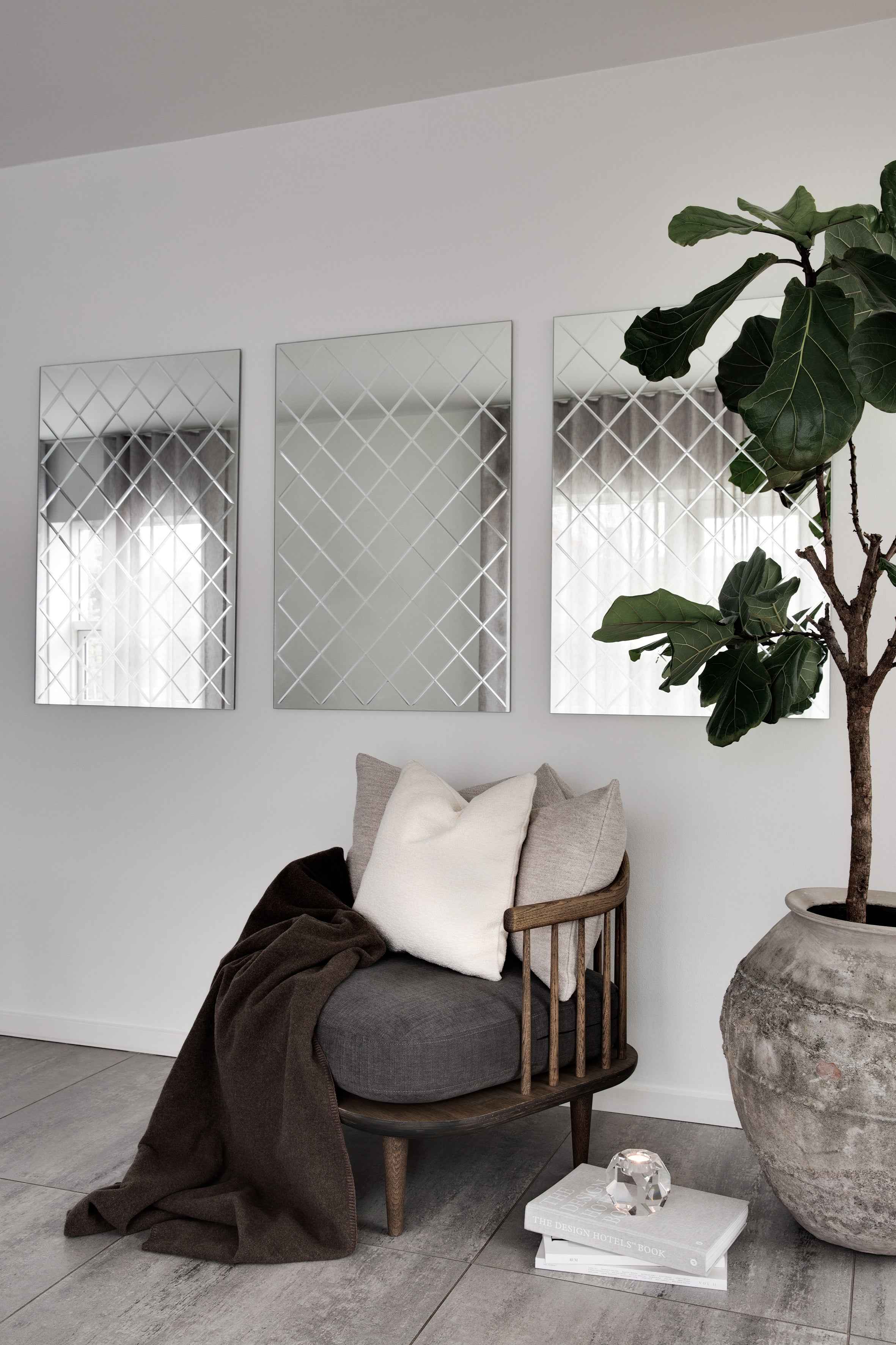 Tre rektangulære spejle med kvadratisk gittermønster hænger side om side på en lys væg. Foran står en stol med puder og plaid, en stabel bøger med en krystal lysestage ovenpå samt en stor grøn plante i krukke.