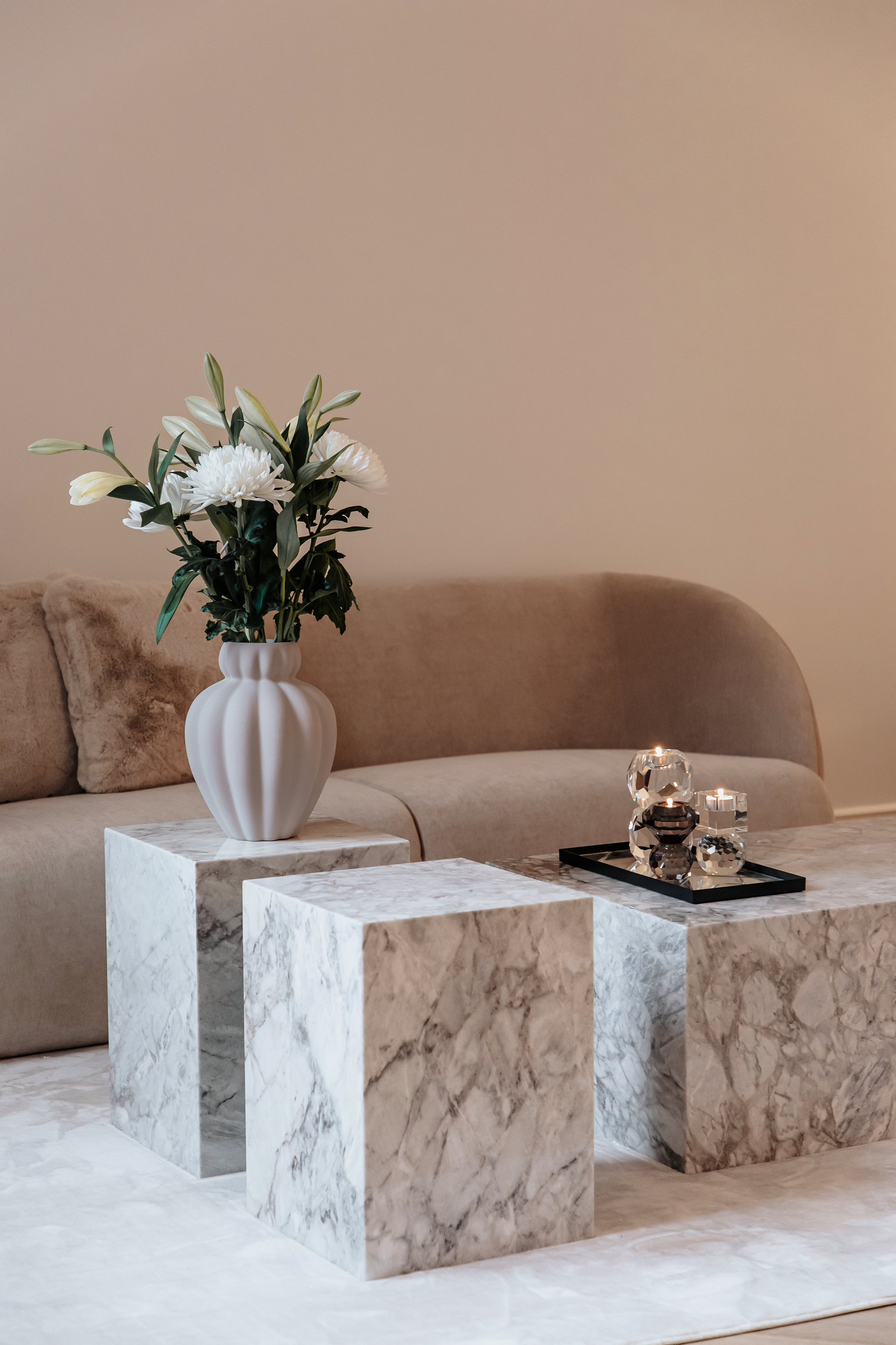 Et interiørfoto af en stue med en lys beige sofa med bløde puder. Foran sofaen står tre kubiske sideborde i hvid- og gråmarmoreret sten. På det ene bord er placeret en hvid, buet vase med hvide blomster og grønne blade. På et andet bord står et sort fad med glas- og metallysestager med tændte lys.