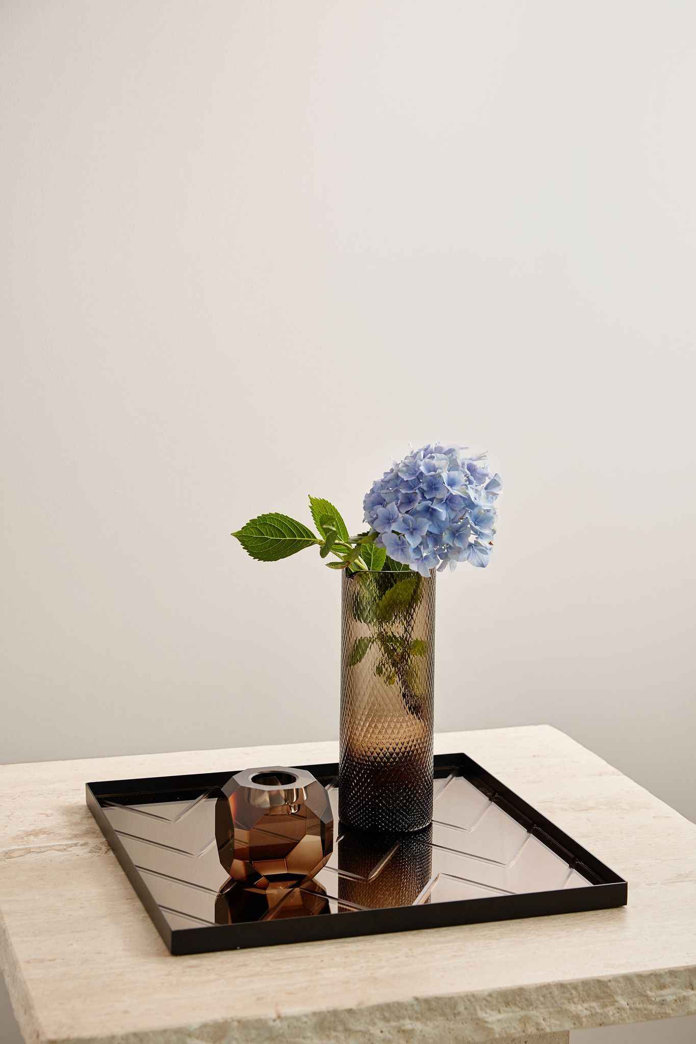Rektangulær spejlbakke med en brun facetslebet fyrfadsstage og en høj brun glasvase med mønster, hvori der står en blå hortensiablomst med grønne blade.