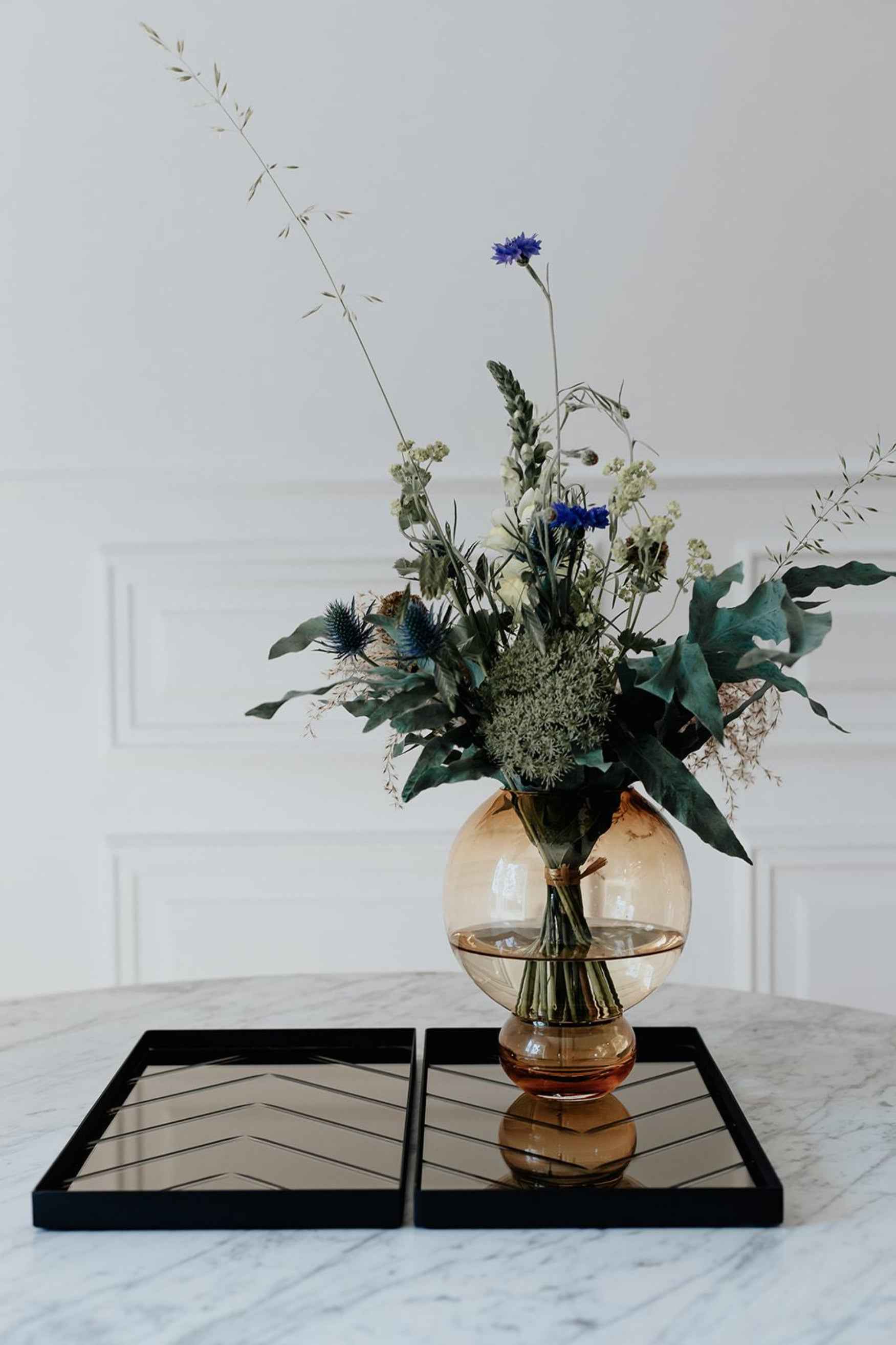 En rund, ravfarvet glasvase med en stor buket blandede blomster og grønne blade står på et marmorbord. Under vasen ligger to rektangulære bakker med spejloverflade placeret side om side.