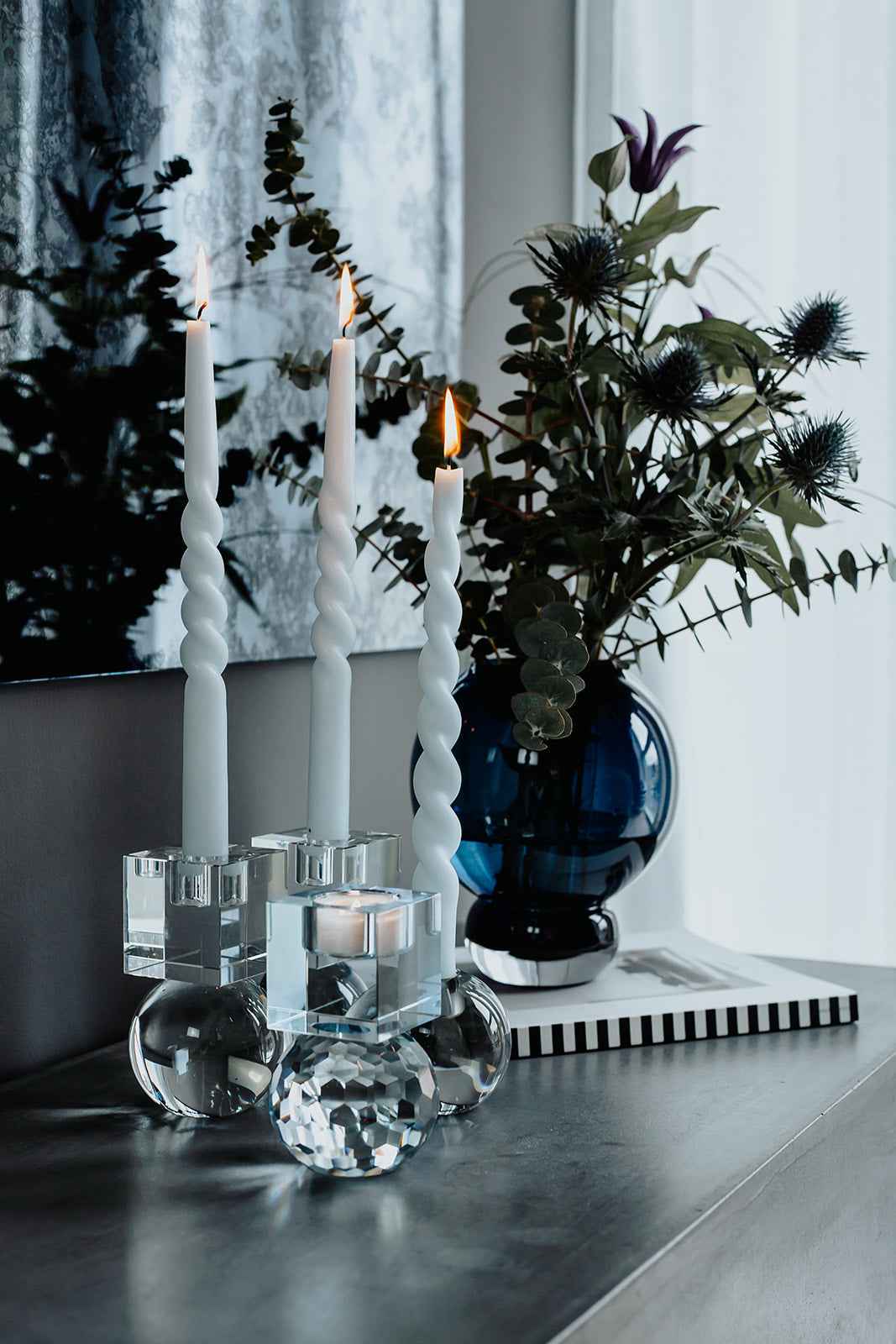 Tre tændte snoede stagelys i farven Mighty Blue placeret i klare glasstager med runde baser, stående på et bord ved siden af en mørkeblå vase med grønne grene og lilla blomster.