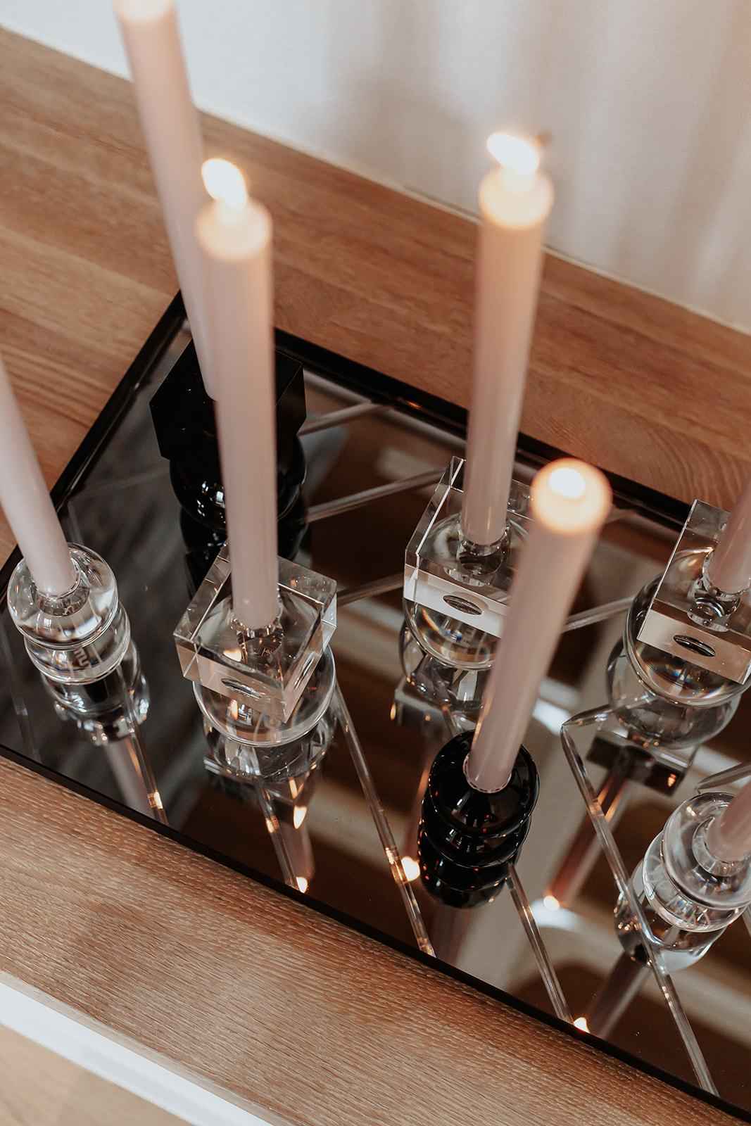 Flere tændte, lyse stearinlys står i forskellige lysestager af glas og sort materiale. Lysestagerne er placeret på en sort, spejlende bakke oven på en træoverflade.