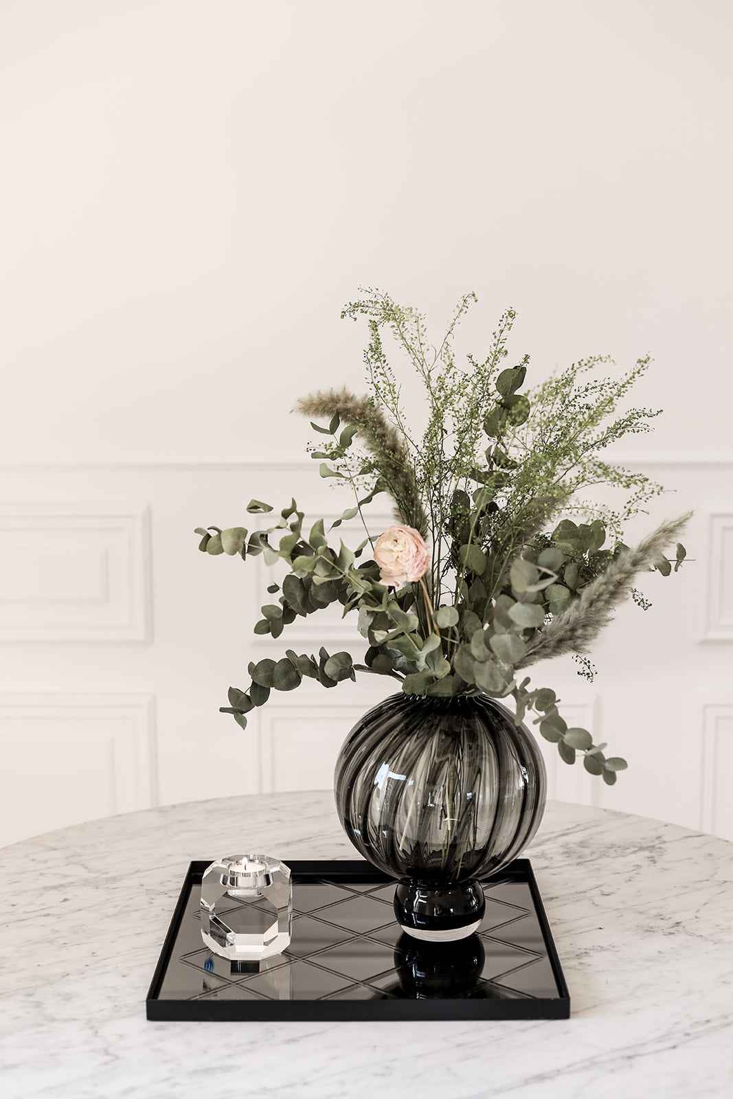 Rundt bord med en sort bakke, dekoreret med en mørk riflet glasvase med grønne grene og en enkelt lys blomst samt en krystal fyrfadsstage.