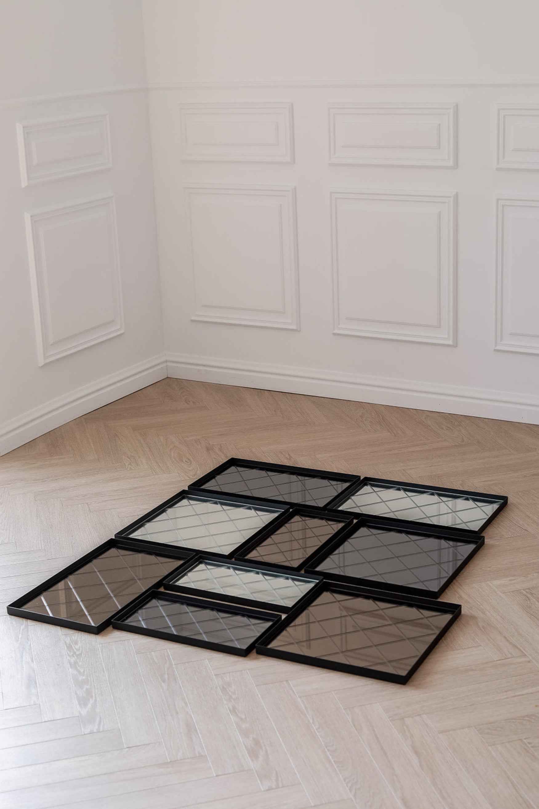 Flere rektangulære bakker med sorte kanter og spejloverflader placeret på et sildebensparketgulv foran en hvid væg med vægpaneler.