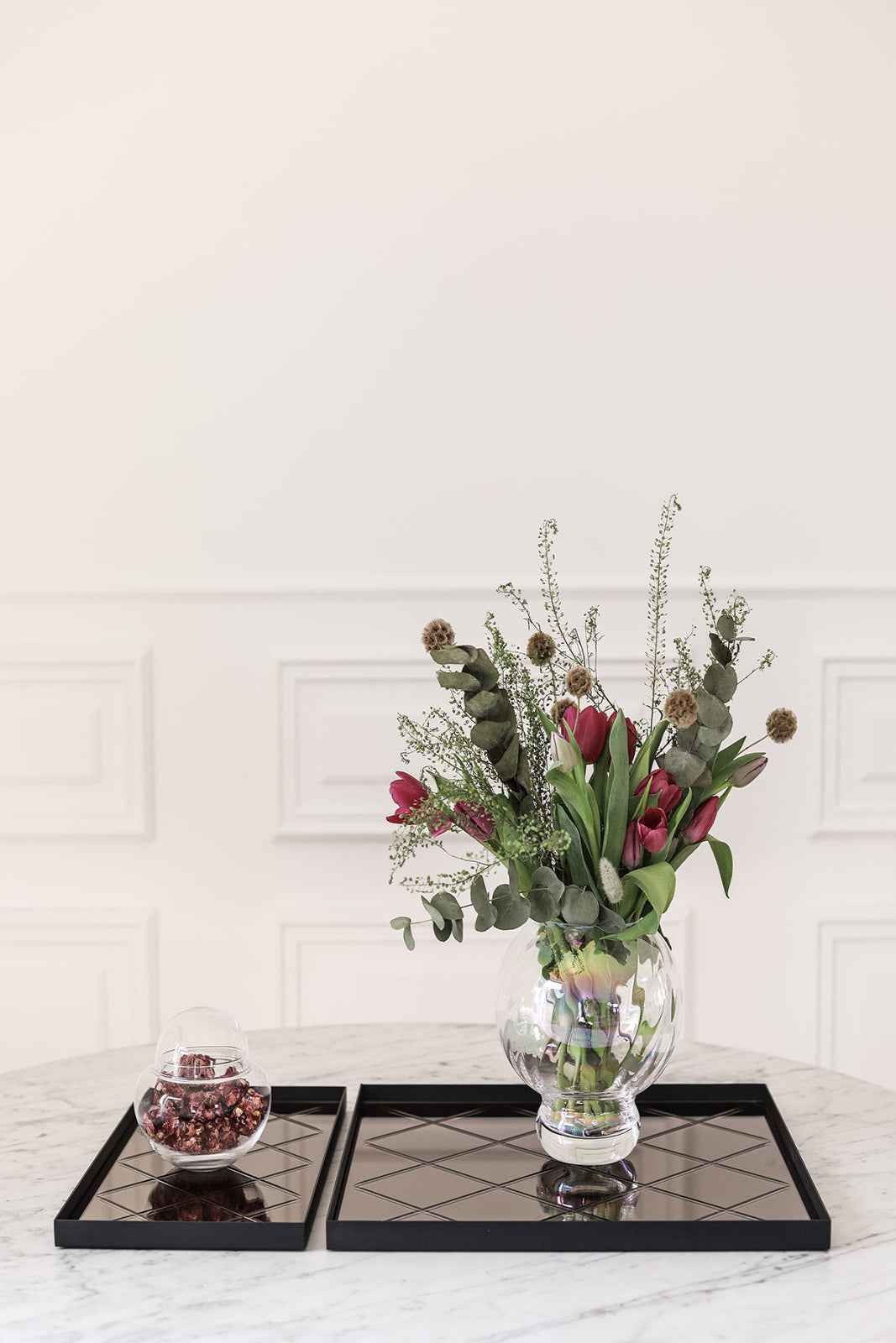 Klar glasvase med røde tulipaner, grønne blade og tørrede blomster placeret på en kvadratisk spejlbakke. Ved siden af står en rund glaskrukke med låg fyldt med tørrede dekorative blomster på en anden spejlbakke.