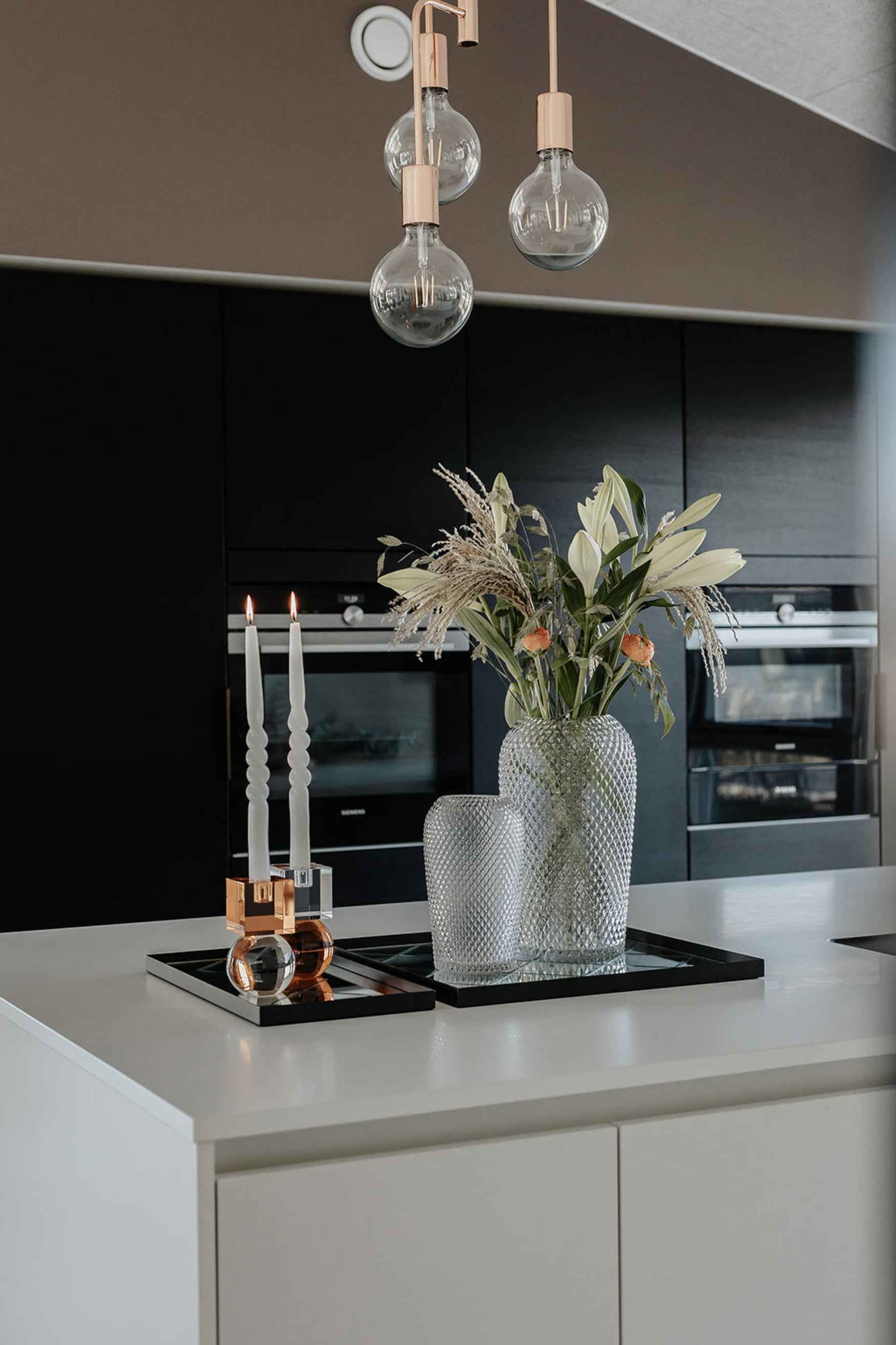 Køkkenø med to glasvaser med mønstret overflade, hvoraf den ene indeholder blomster, placeret på sorte spejlbakker. Ved siden af står to tændte stearinlys i moderne lysestager.