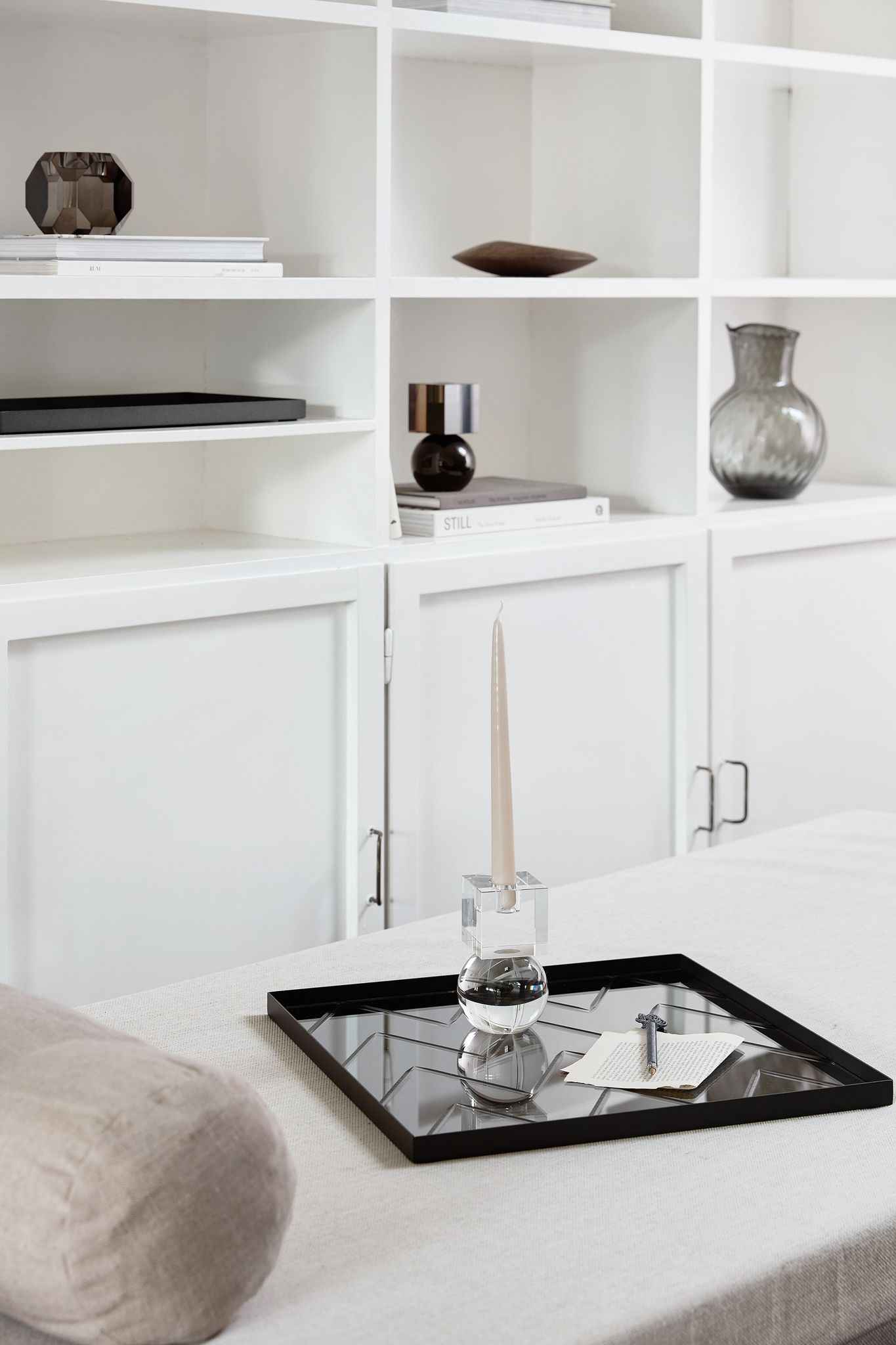 Sort firkantet spejlbakke med geometrisk mønster placeret på en lys overflade; på bakken står en høj, slank lys beige stearinlys i en klar krystal-lysestage, ved siden af et stykke papir med tekst og en kuglepen. I baggrunden ses en hvid reol med forskellige dekorative objekter, herunder glasvaser og bøger.