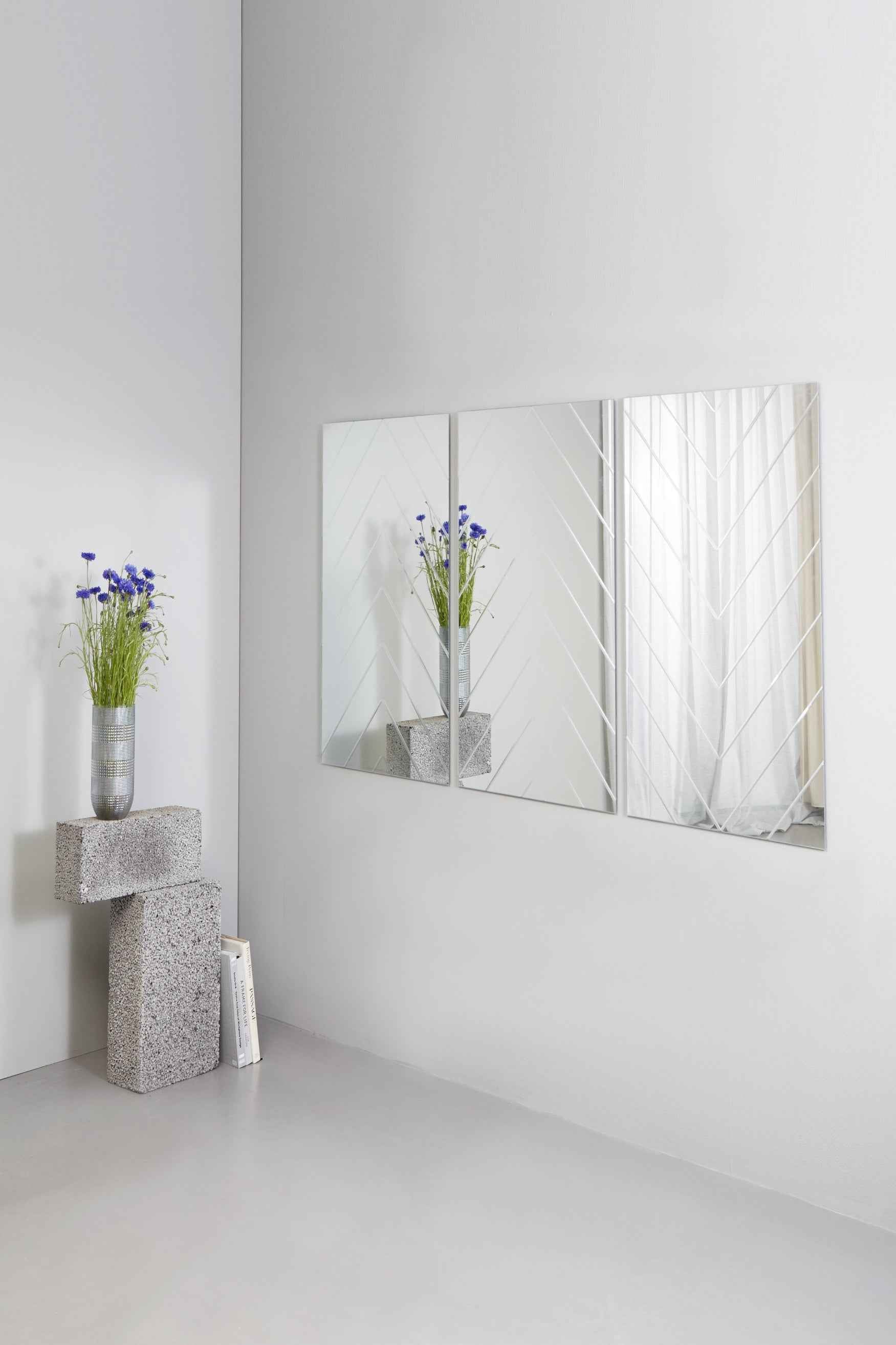 Tre rektangulære spejle med sildebensmønster hænger side om side på en lys væg. Ved siden af står en vase med blå blomster oven på en skulpturel piedestal af grå stenblokke.
