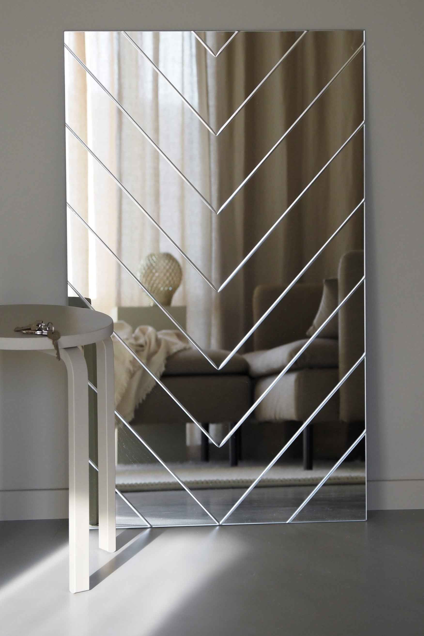 Rektangulært spejl med sildebensmønster står op ad en væg og spejler en sofa, gardiner og en lampe. Til venstre ses et lille bord med en nøgle på kanten.