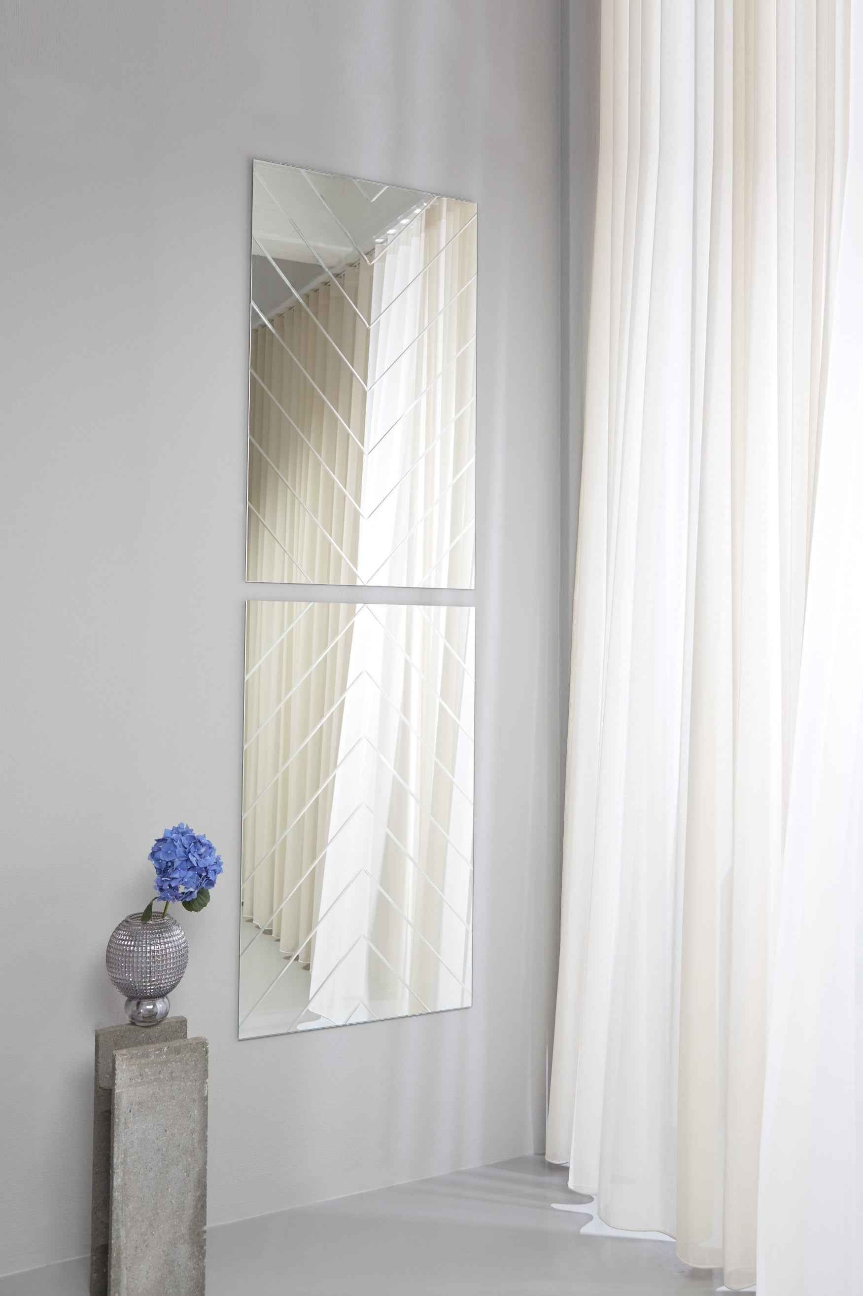 To rektangulære spejle med sildebensmønster hænger lodret over hinanden på en lys væg ved siden af hvide gardiner. Foran står en piedestal i sten med en rund vase med en blå blomst.