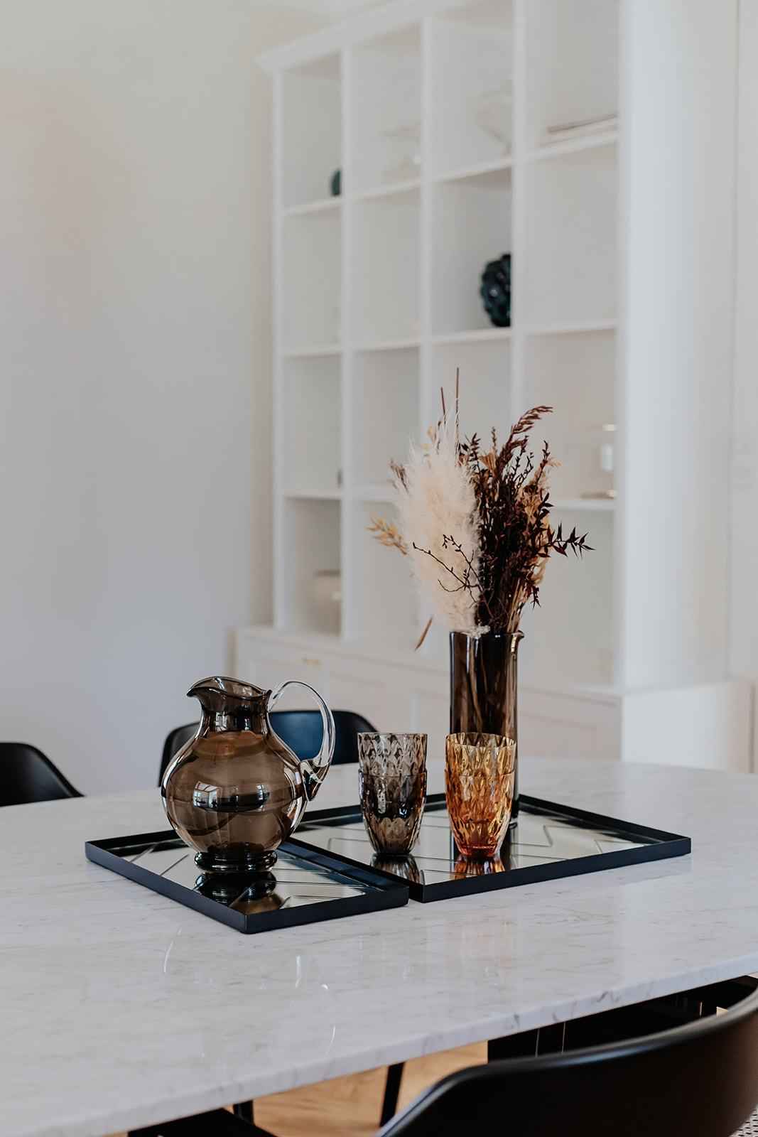 To sorte firkantede spejlbakker med diagonalt mønster placeret på et marmorbord; på bakkerne står en brun glaskande, to facetslebne drikkeglas i ravgul og brun samt en høj brun glasvase med tørrede grene og pampasgræs.