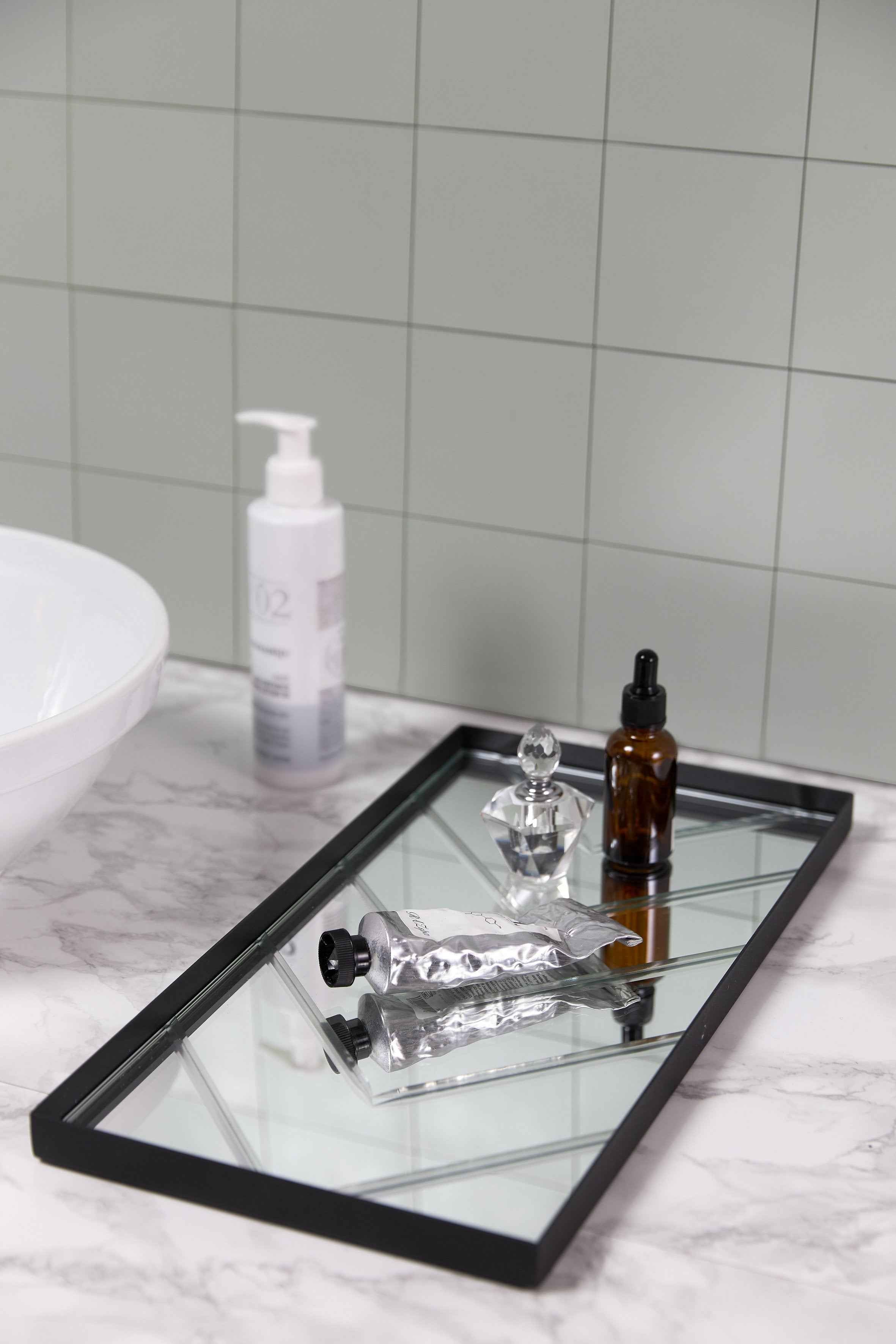 En sort bakke med spejloverflade står på en marmorlignende bordplade. På bakken ligger en sølvfarvet tube samt en lille brun glasflaske med pipette og en klar parfume- eller glasflakon med slebet låg. I baggrunden ses en pumpeflaske og en hvid håndvask.