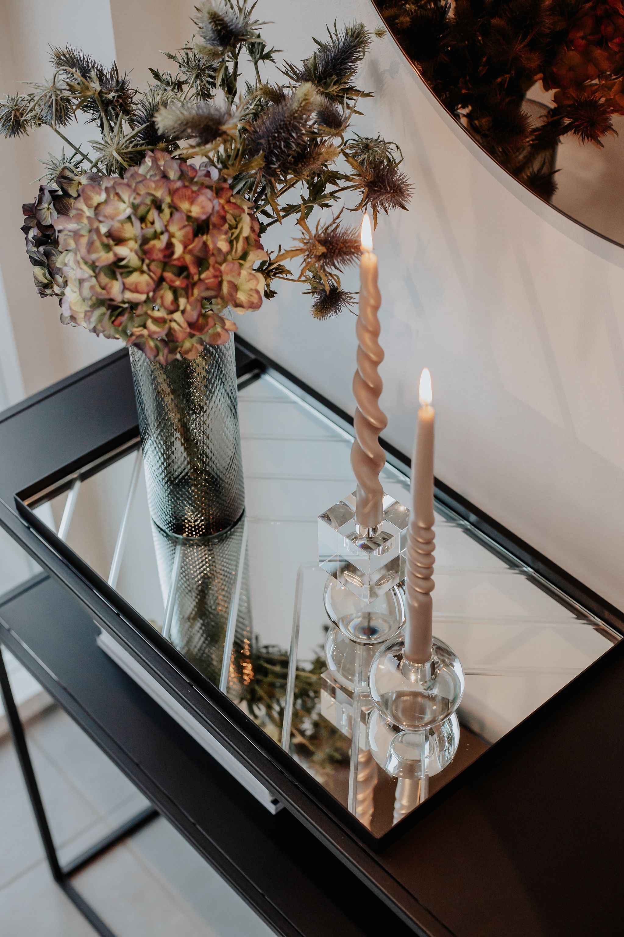 ChatGPT sagde:
En glasvase med tørrede blomster står på en sort bakke med spejloverflade. Ved siden af vasen er to tændte, snoede stearinlys placeret i klare glaslysestager.