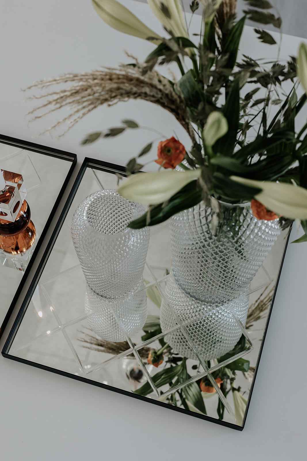 Nærbillede af to sorte rektangulære bakker med spejloverflade, dekoreret med en riflet glasvase med blomsterarrangement og en gennemsigtig fyrfadsstage.