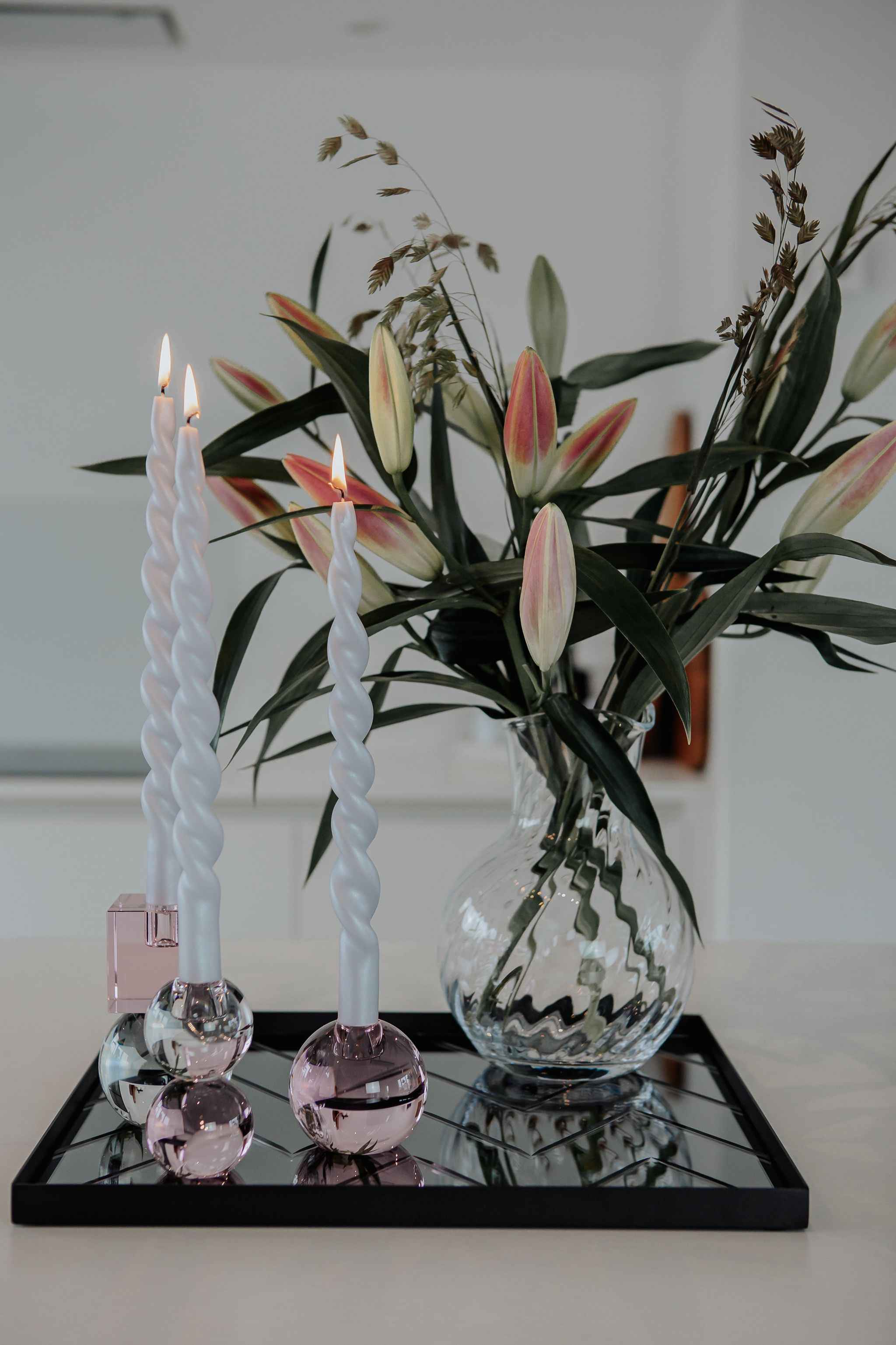 Sort firkantet spejlbakke med diagonalt mønster placeret på en lys bordplade; på bakken står to tændte snoede hvide stearinlys i lysestager med kugleformet fod i rosa og klar glas. Ved siden af står en stor gennemsigtig glasvase med et bundt lyserøde liljeknopper og grønne blade.