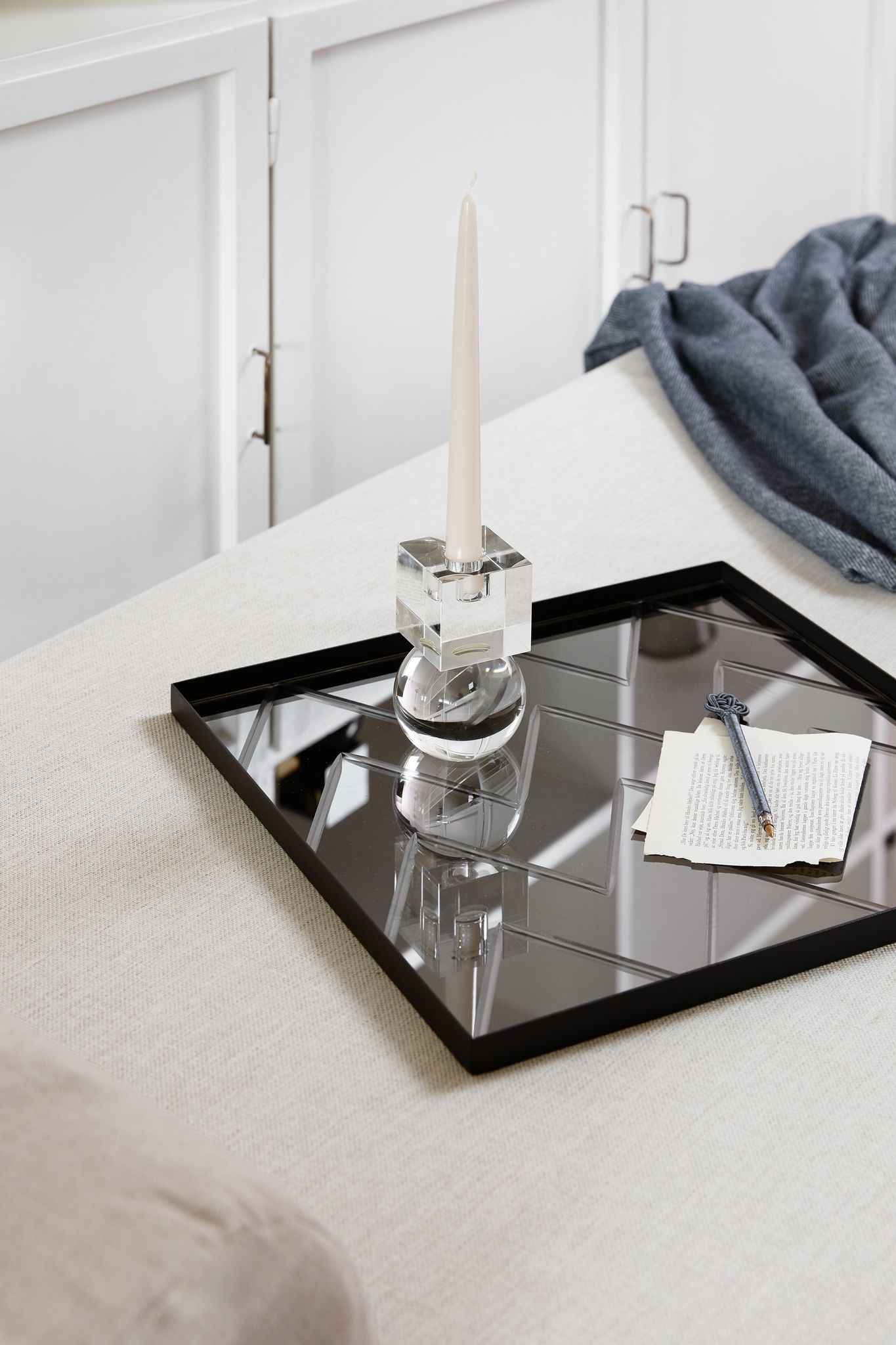 Sort firkantet spejlbakke med geometrisk mønster placeret på en lys overflade; på bakken står et højt, slankt, beige stearinlys i en klar krystal-lysestage, og ved siden af ligger et stykke papir med tekst samt en dekorativ pen. I baggrunden ses et mørkeblåt tekstil over møblet.