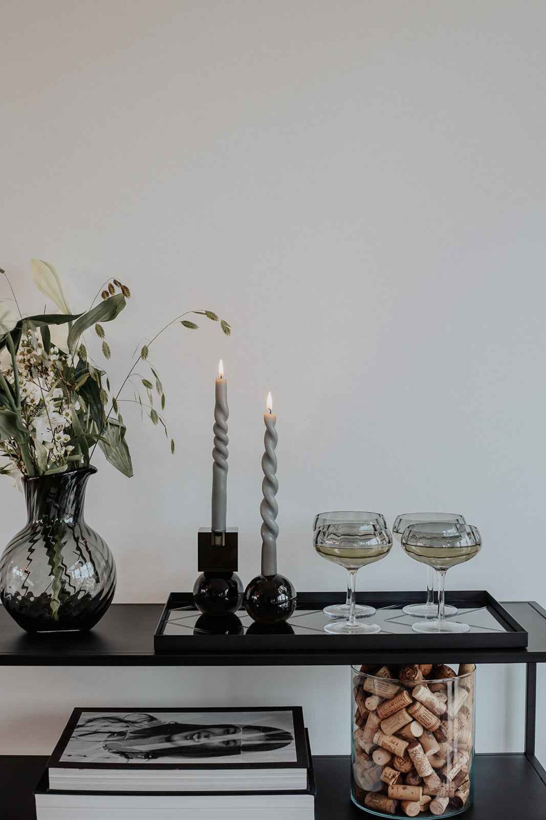Et sort konsolbord dekoreret med en glasvase med grønne blade og hvide blomster, to tændte snoede grå stearinlys i mørke lysestager samt fire fyldte champagneglas. På nederste hylde ligger store bøger, og et glas er fyldt med vinpropper.
