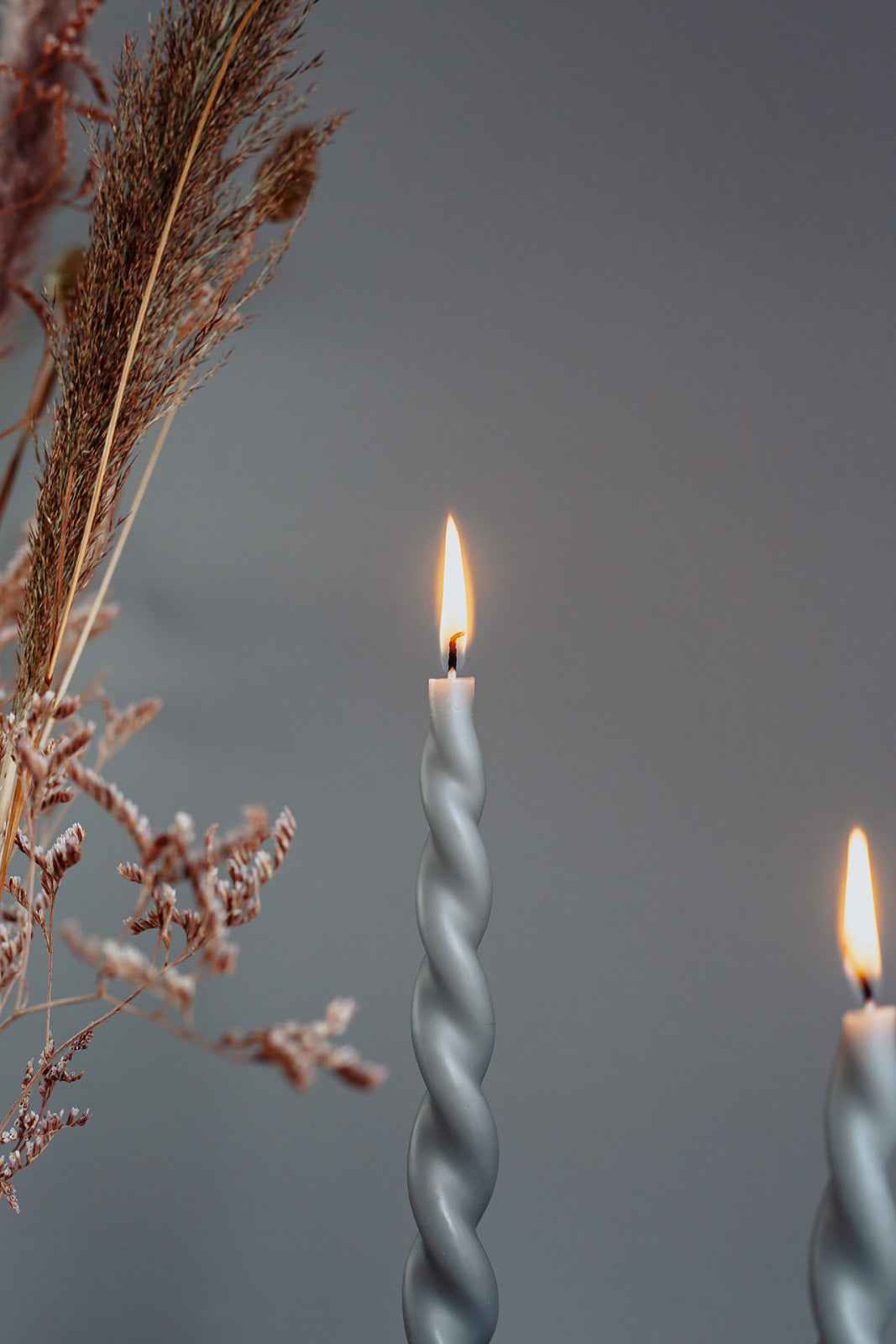 Nærbillede af en tændt snoet grå stearinlys med flamme, omgivet af tørrede græsser og kviste mod en grå baggrund.