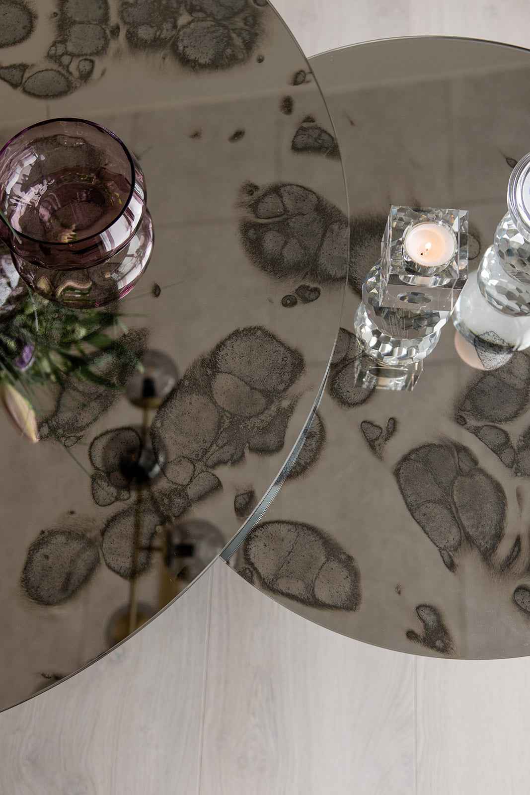 Nærbillede af to runde sofaborde med mønstret spejlglasplade, dekoreret med en lilla glasskål og en krystal fyrfadsstage med tændt lys.