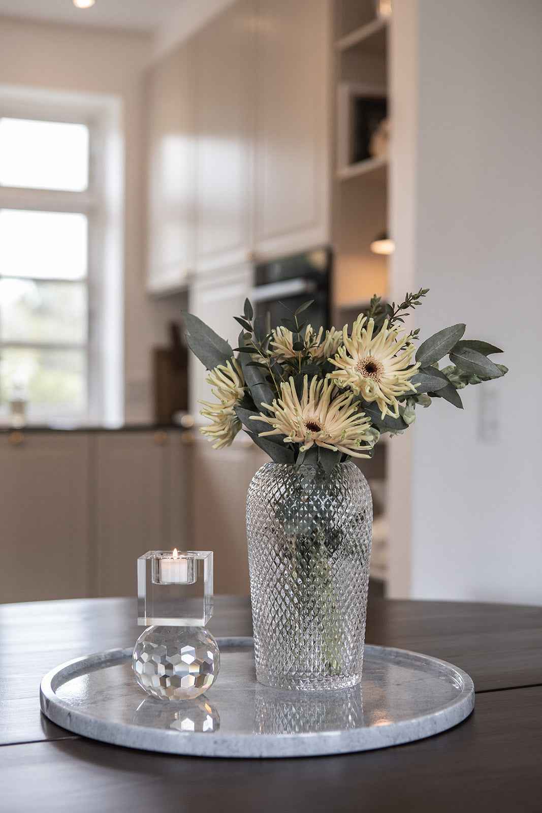 Rund bakke i grå marmor placeret på et mørkt bord, dekoreret med en gennemsigtig rillet glasvase med blomster og en krystal fyrfadsstage med lys.