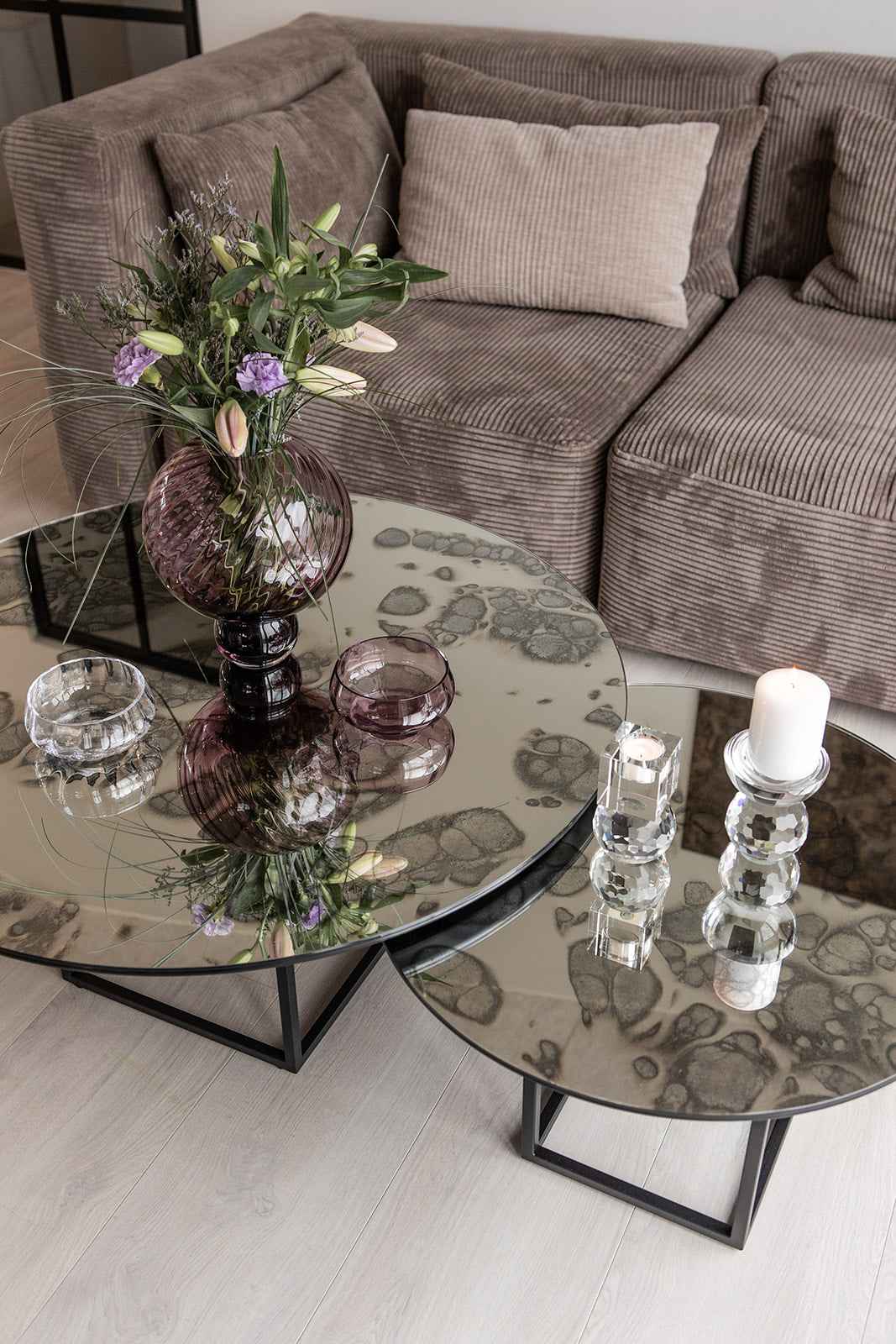 "To runde sofaborde med mønstret spejlglasplader og sorte metalben, dekoreret med en lilla glasvase med blomster, små glasskåle og en krystal lysestage med hvidt stearinlys, placeret foran en brun fløjlssofa med puder.