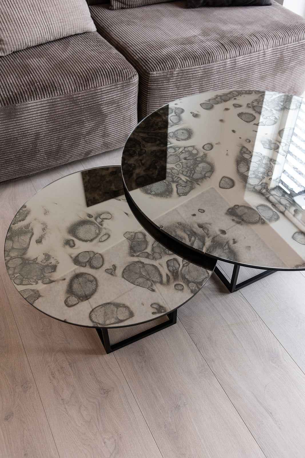 To runde sofaborde i forskellig højde med spejlglasplader med mønster og sorte metalben, placeret foran en brun fløjlssofa.