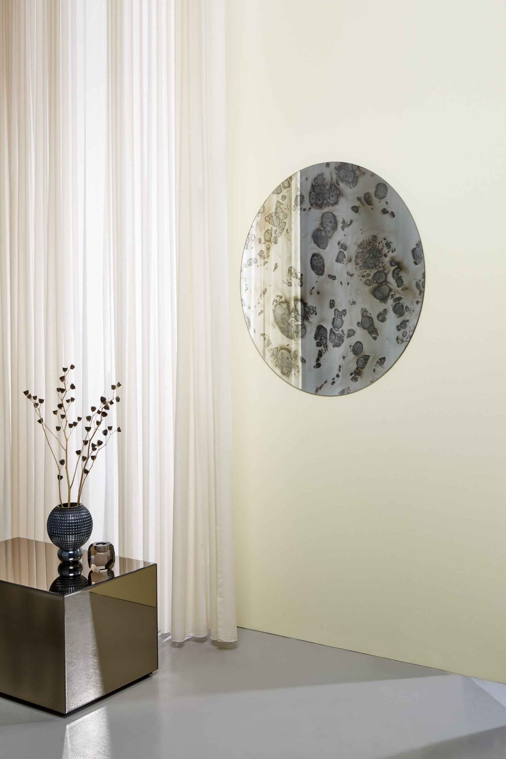 Rundt vægspejl med antikt mønster hænger på en lys væg ved siden af hvide gardiner. Foran står et lille bord med spejlende overflade, dekoreret med en blå vase med tørrede grene og en lille glasdekoration.