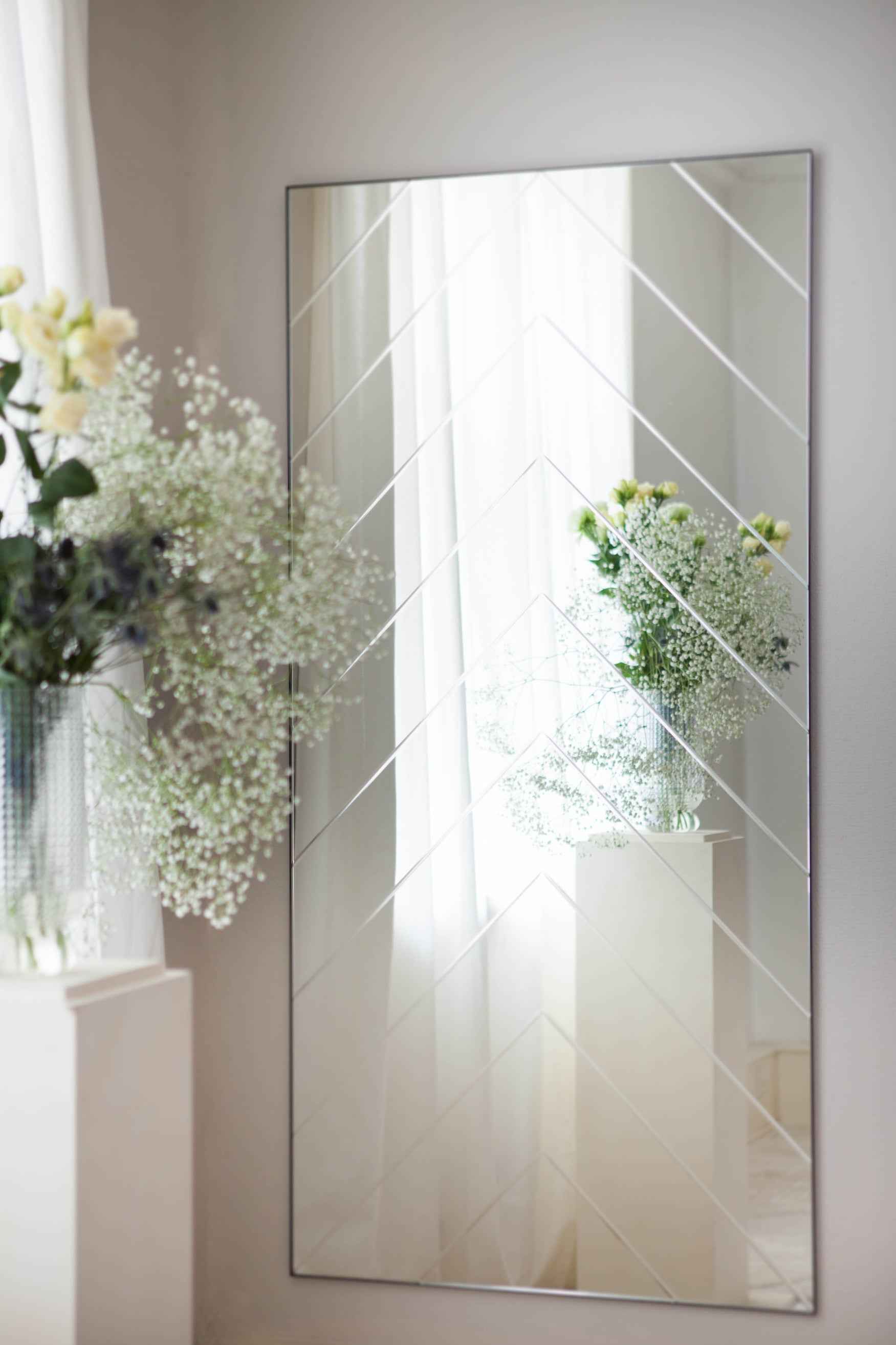 Rektangulært vægspejl med chevron-mønster hænger på en væg, hvor det spejler hvide gardiner og en glasvase med blomster og grønt.