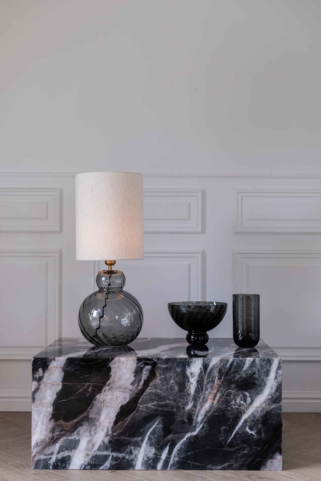 Et marmorbord i sort og hvid marmor står foran en væg med klassiske paneler. På bordet står en bordlampe med rund fod i røgfarvet glas, messingdetalje og en høj, cylinderformet hvid lampeskærm. Ved siden af lampen er placeret en sort glasskål på fod og et højt røgfarvet glas. Gulvet er i lyst sildebensparket.