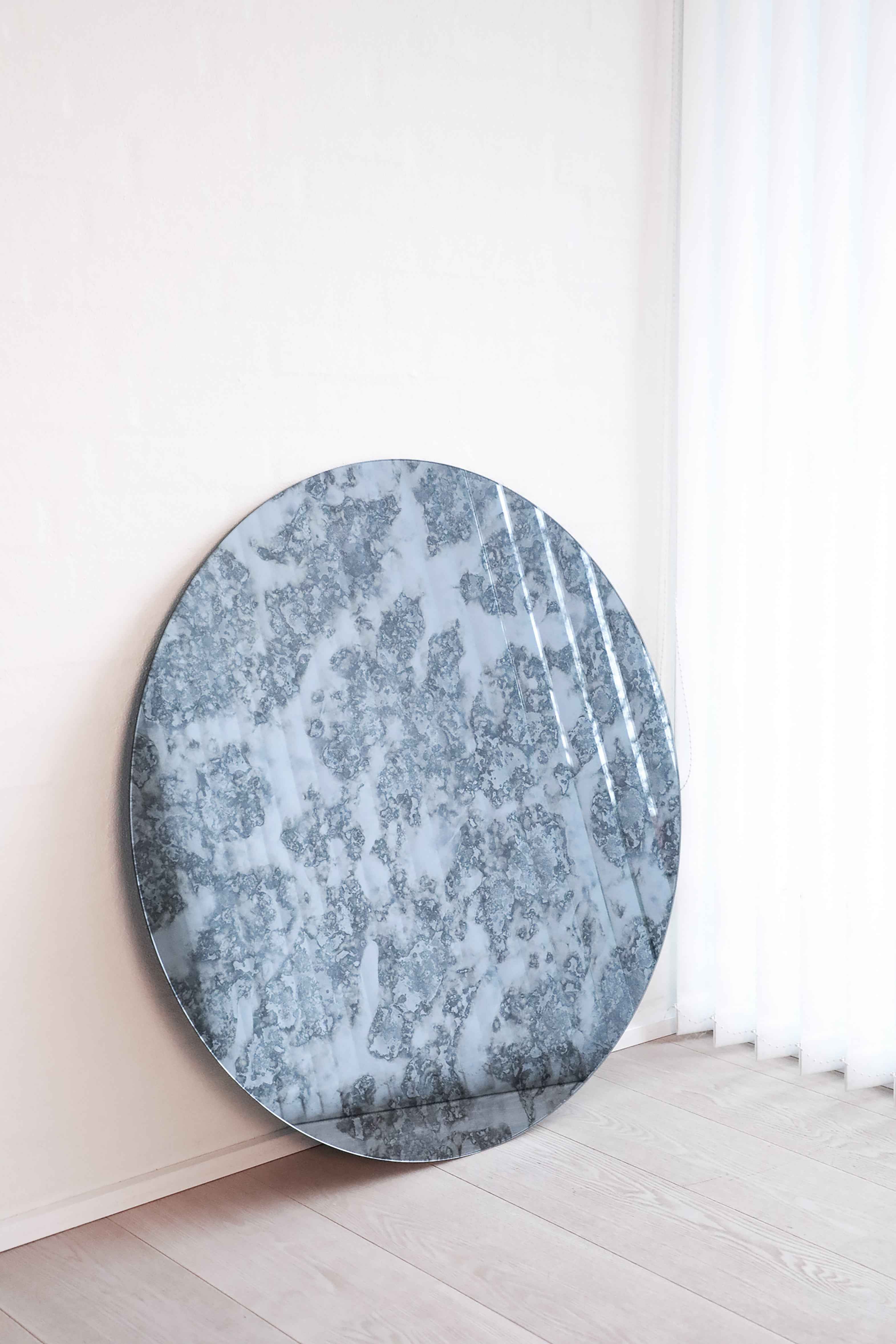 Rundt spejl med gråblåt antikt mønster lænet op ad en lys væg på et gulv i lyst træ, ved siden af et vindue med hvide gardiner.