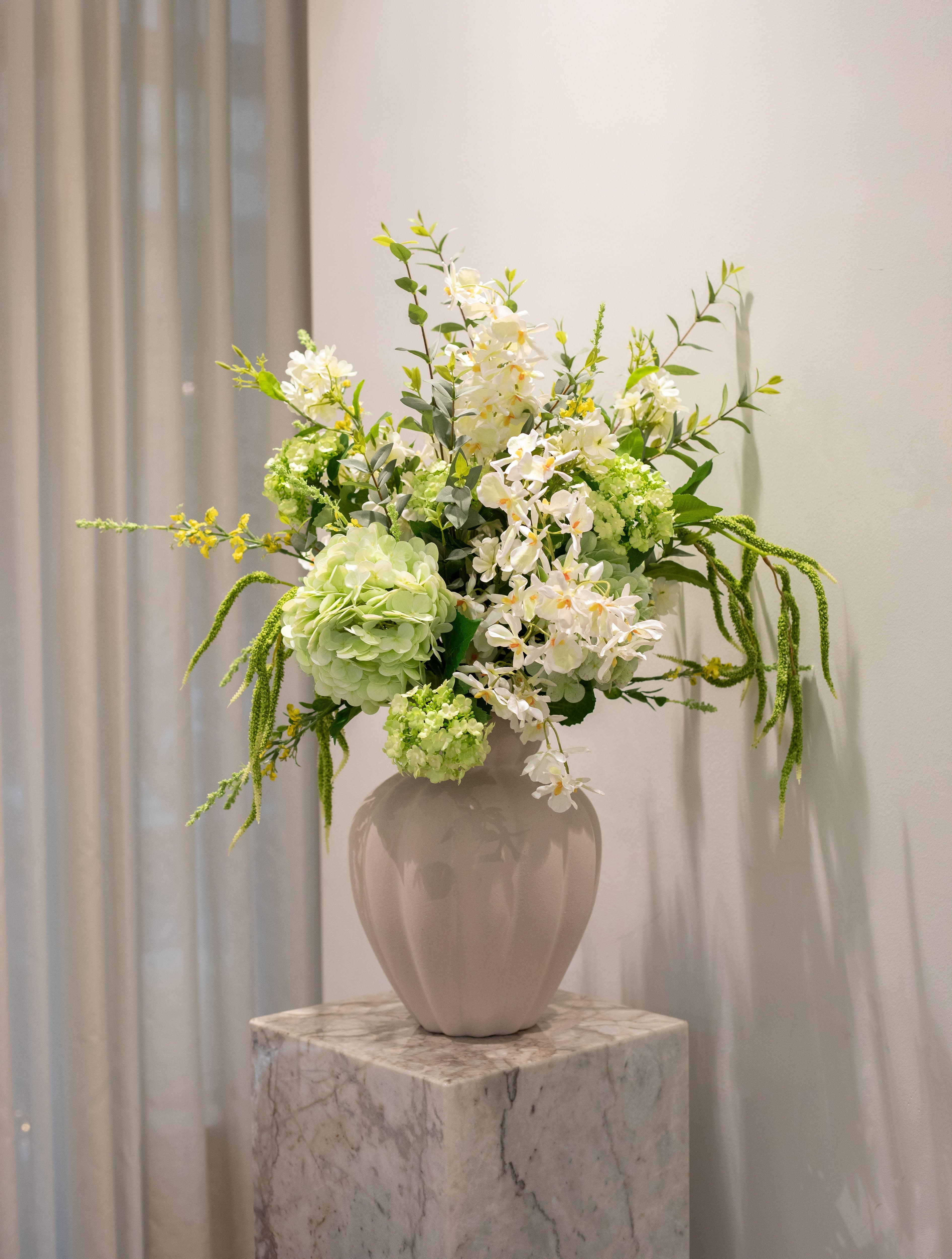 Billede af Specktrum vase i beige, der står på en marmor pedestal. I vasen er en buket kunstige blomster i hvide og grønne nuancer.