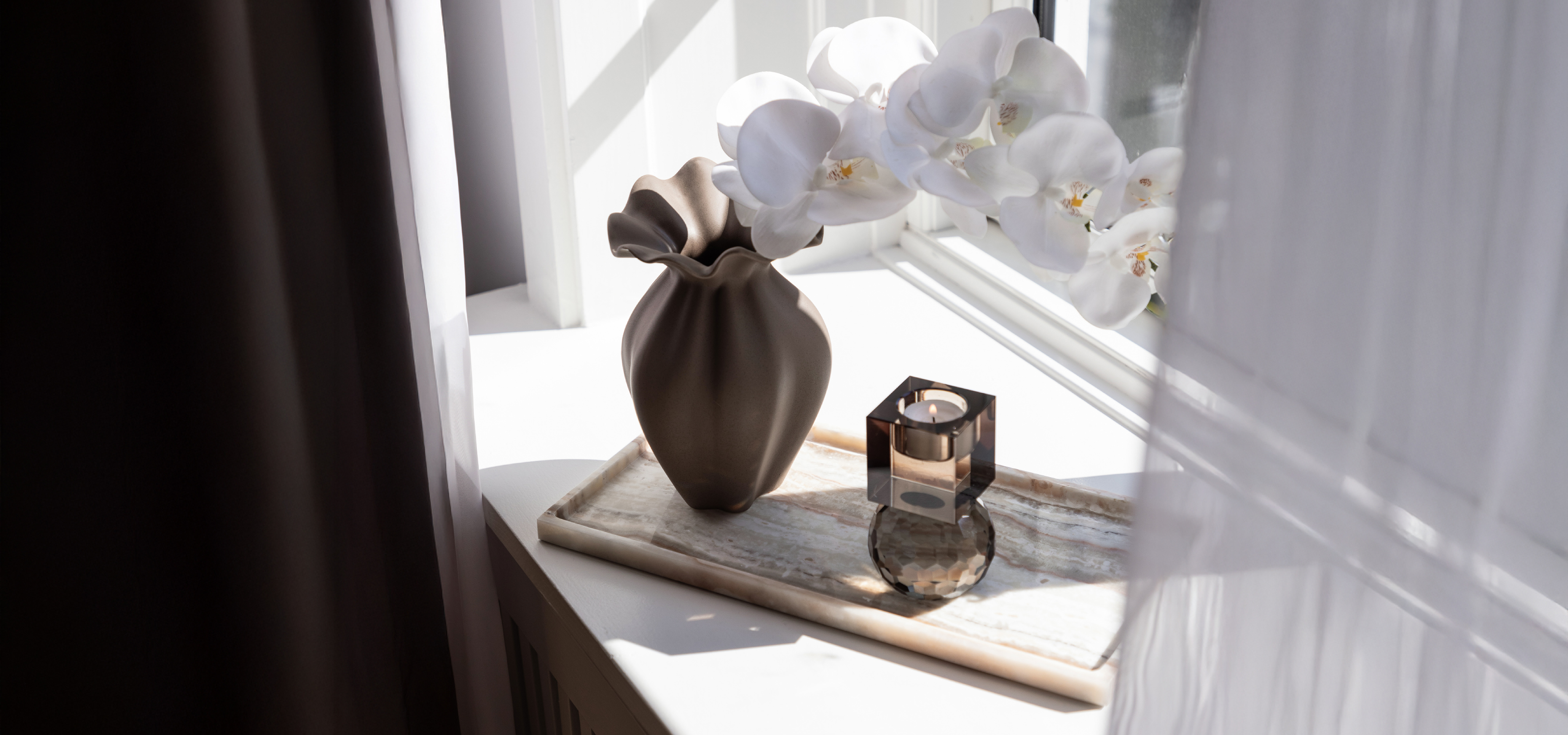 Hvid marmorbakke placeret i en vindueskarm. På bakken er der placeret en brun keramikvase med en hvid orkidé i, ved siden af vasen ses en mørk krystallysestage til fyrfadslys.