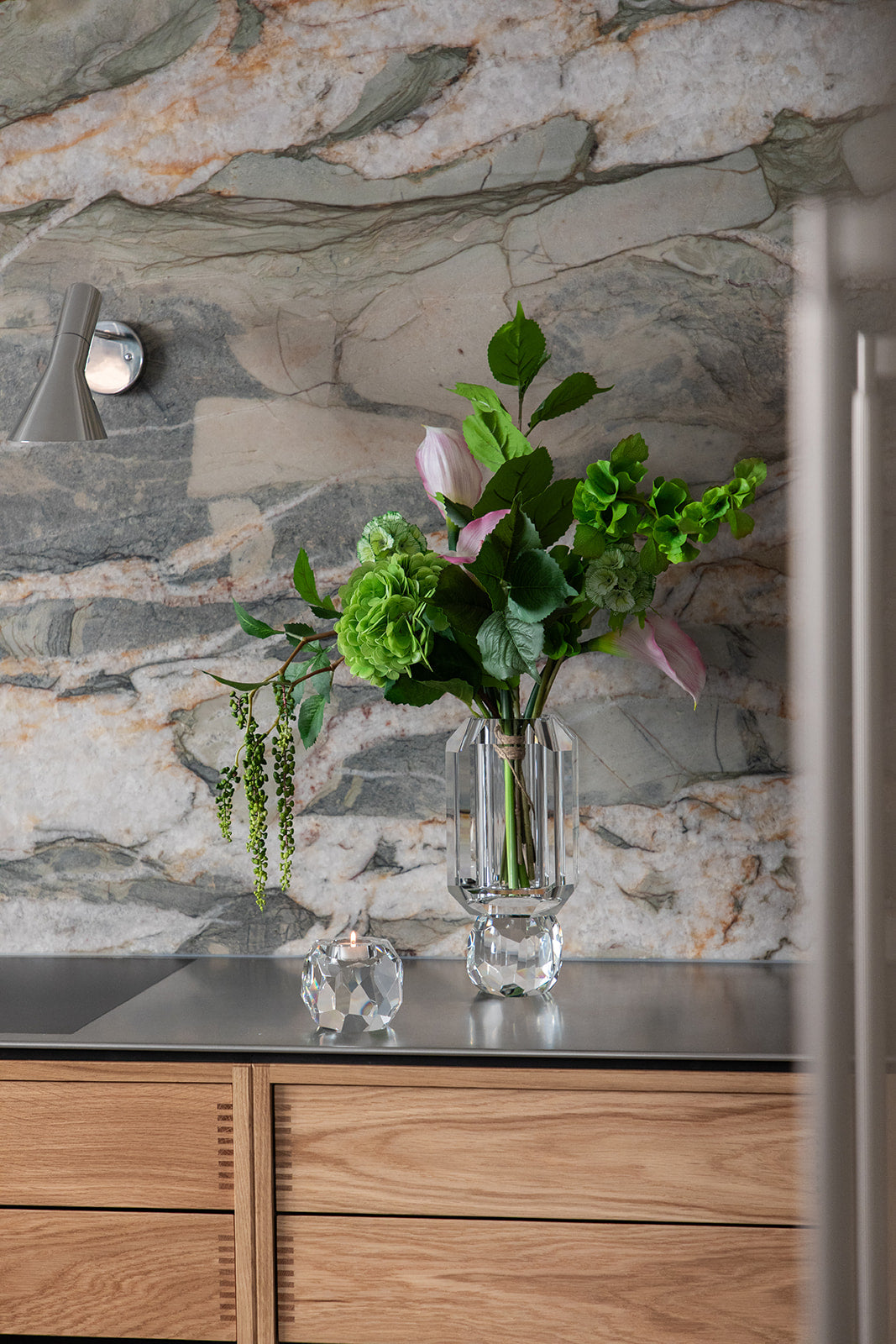Gennemsigtig krystal vase, der står på et mørkt træ bord. I vasen er en buket kunstige blomster i lyse nuancer.
