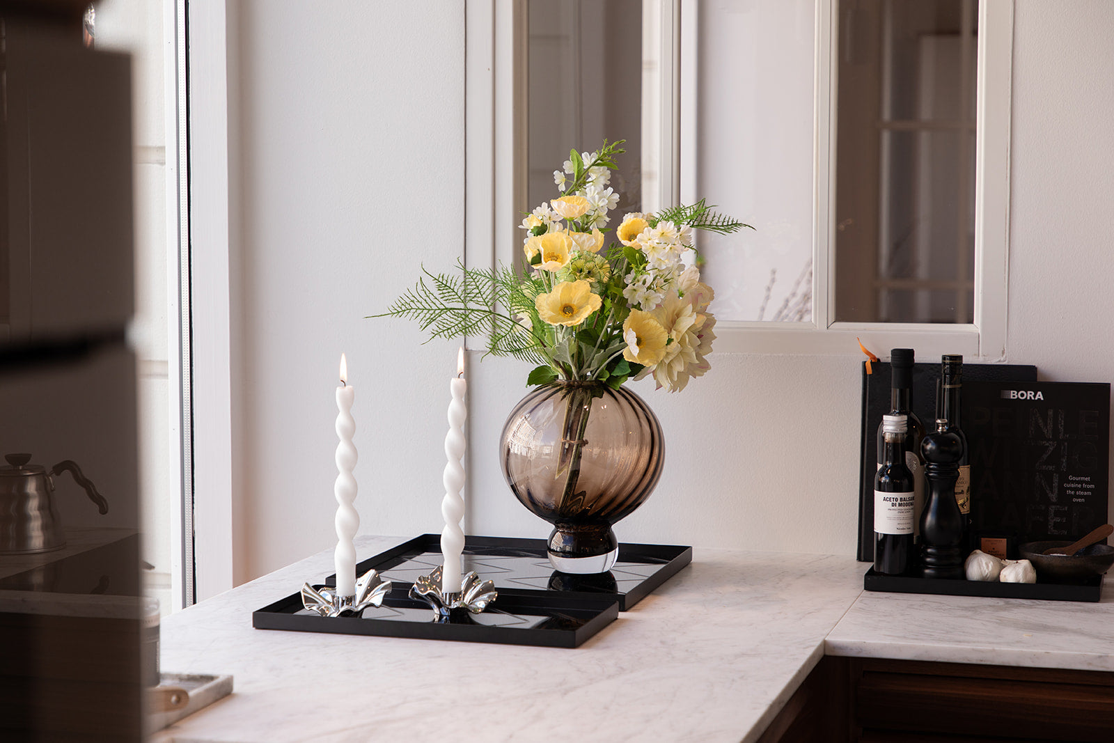 En rund brun glas vase, med kunstige blomster, der står på en spejlbakke. Ved siden af er endnu en spejlbakke med lysestager og snoede lys. Står på et køkkenbord. 