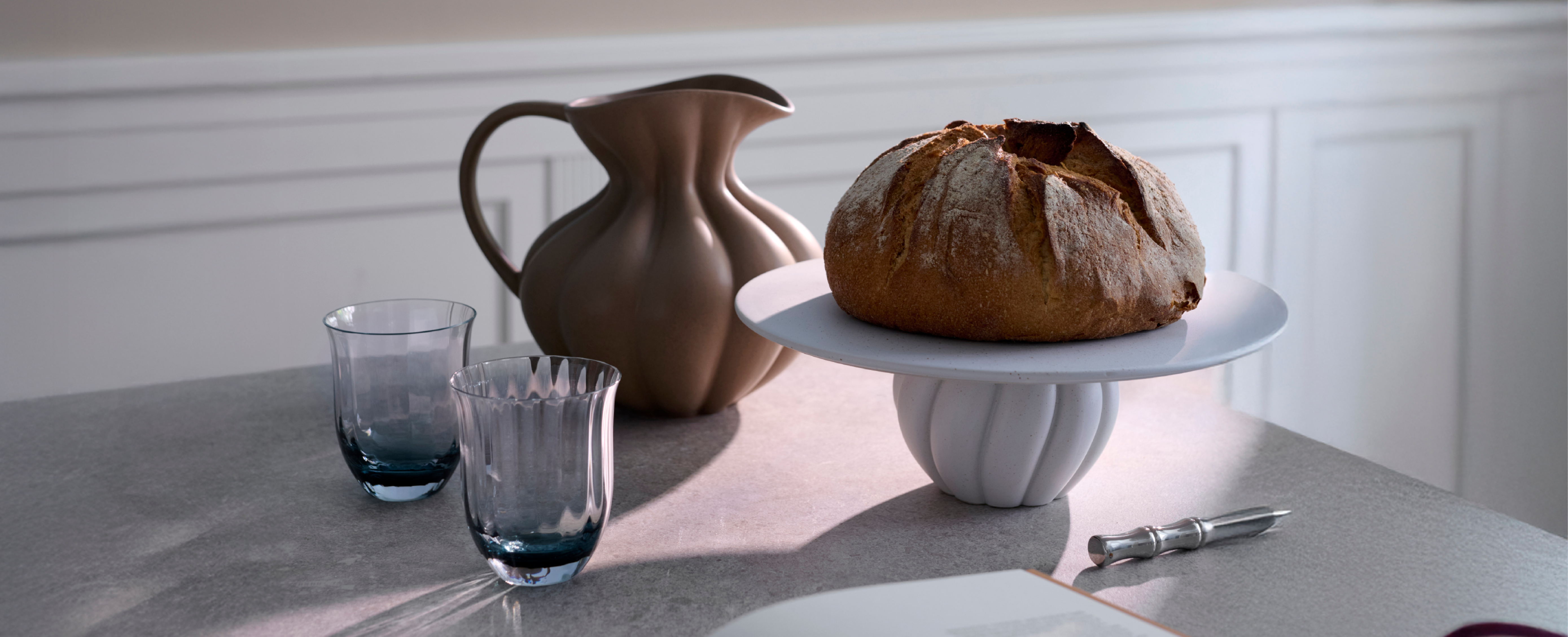 Et spisebord hvorpå der står et keramik opsatsfad med et brød, en keramik kande og to drikkeglas i glas.