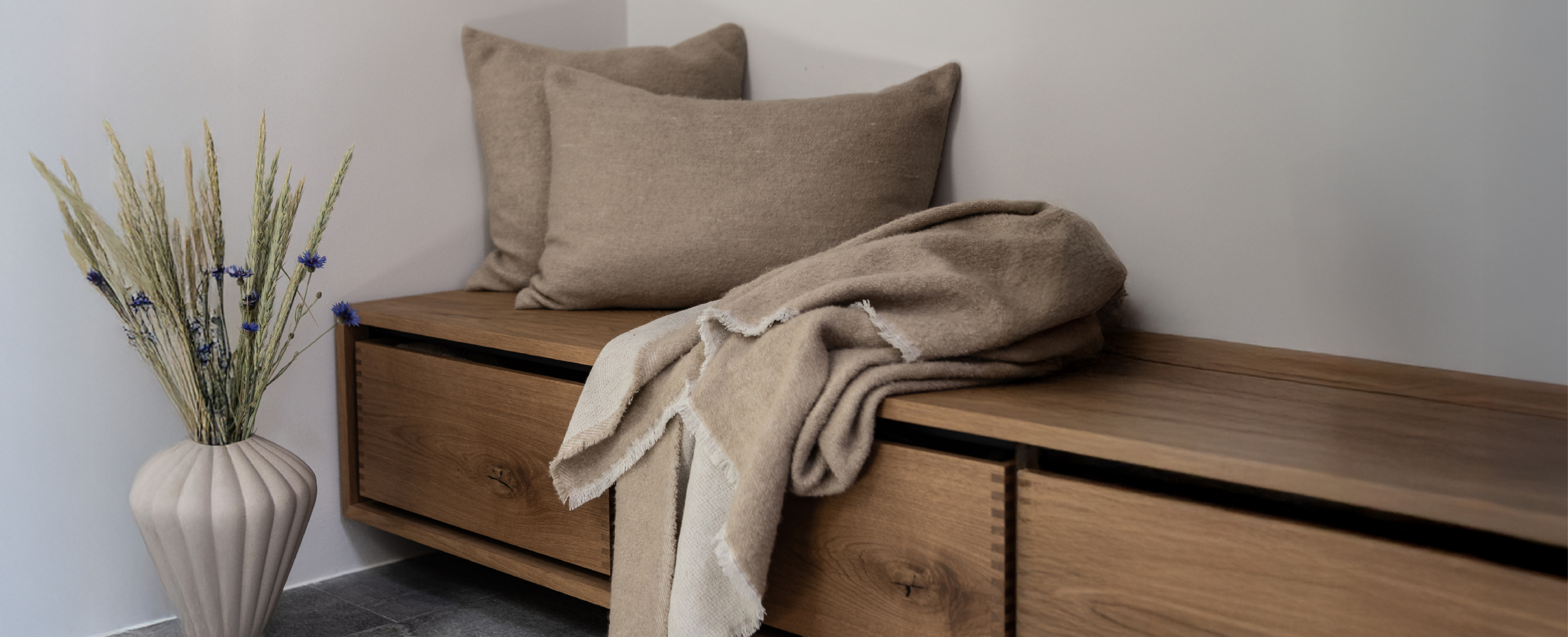 Brun træ bænk i en entre, ovenpå er to beige puder og en beige plaid. På gulvet står en brun keramikvase med små siv og blomster i.