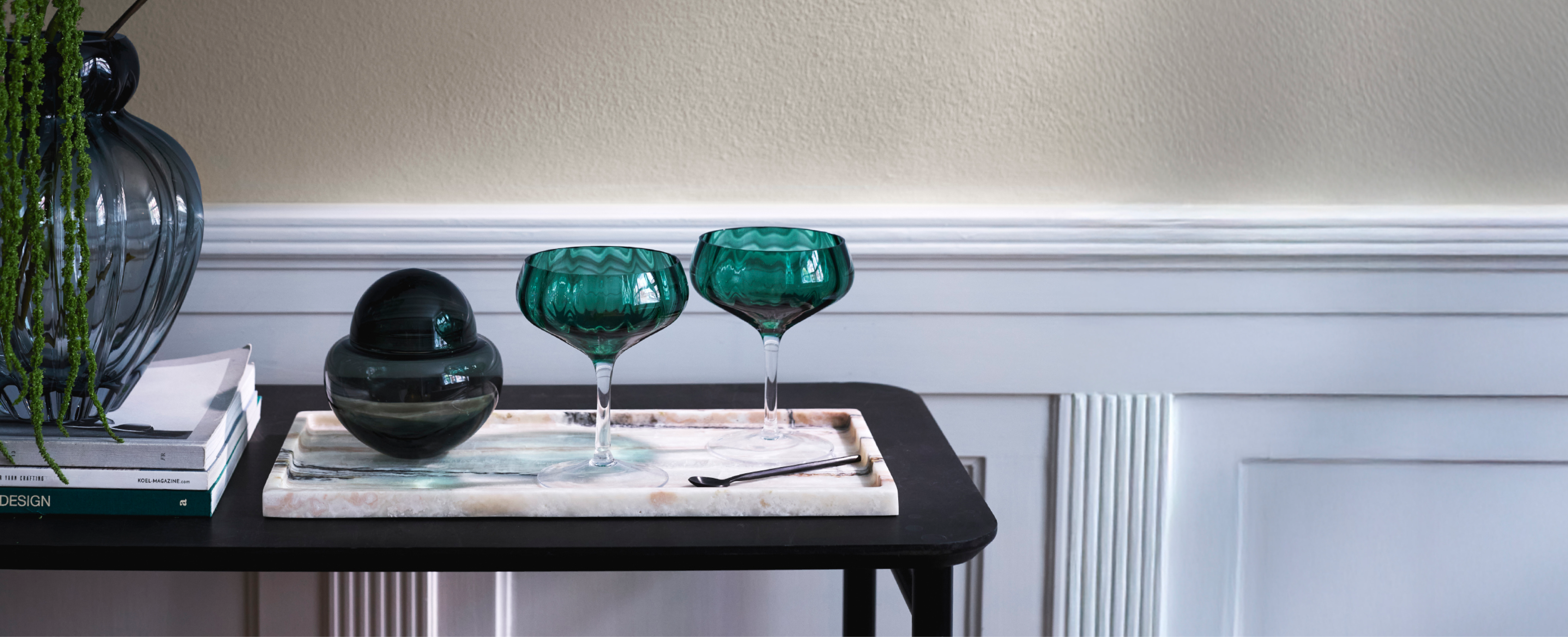 To grønne cocktail glas, der står på en marmorbakke med en matchende bonbonniere. Ved siden af ligger magasiner og oven på dem står en matchende glasvase. Det er hele er sat ovenpå en sort reol.