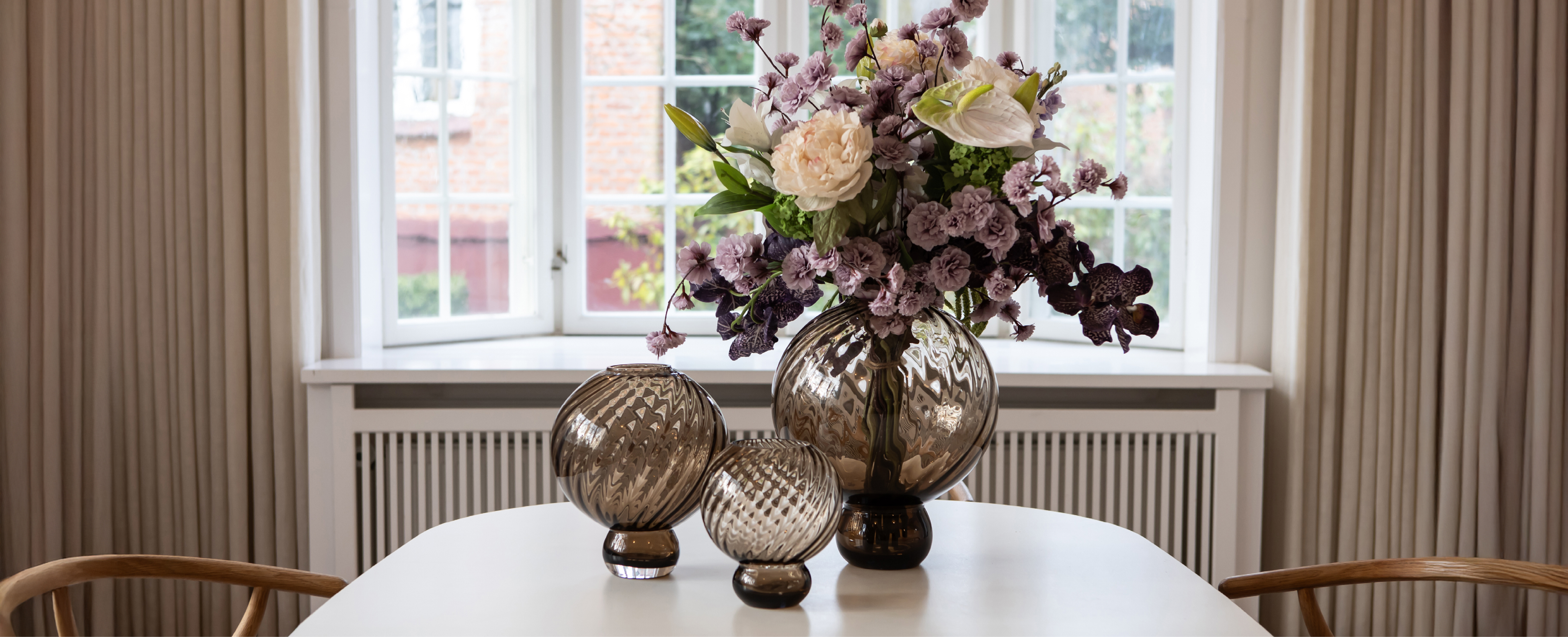 Billede af en spisestue, i baggrunden er et stort vindue med lange beige gardiner. På spisebordet står tre Meadow glas vaser i farven brun fra Specktrum. I den store vaser er der placeret en buket kunstige blomster i hvide, lilla og grønne nuancer.