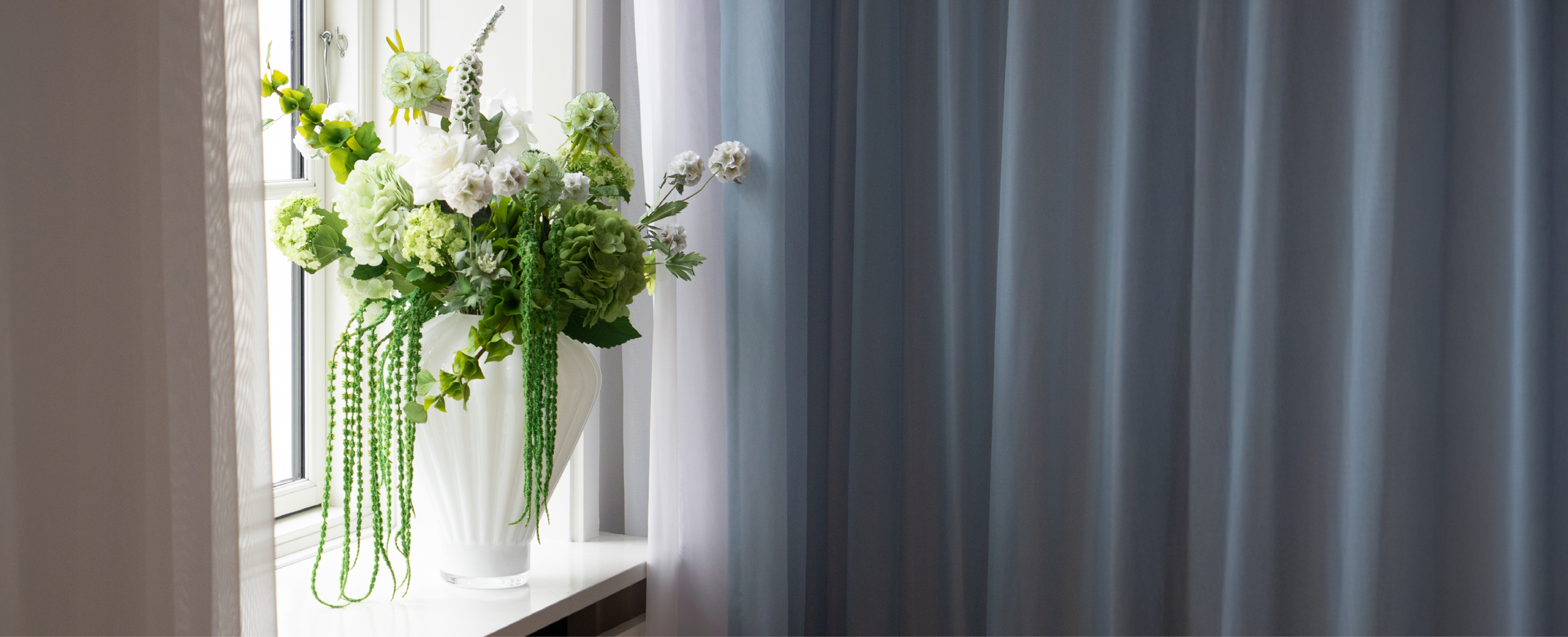 En grøn buket kunstige bloster, der står i en hvid glas vase i en vindueskarm. Baggrunden er et blåt gardin.