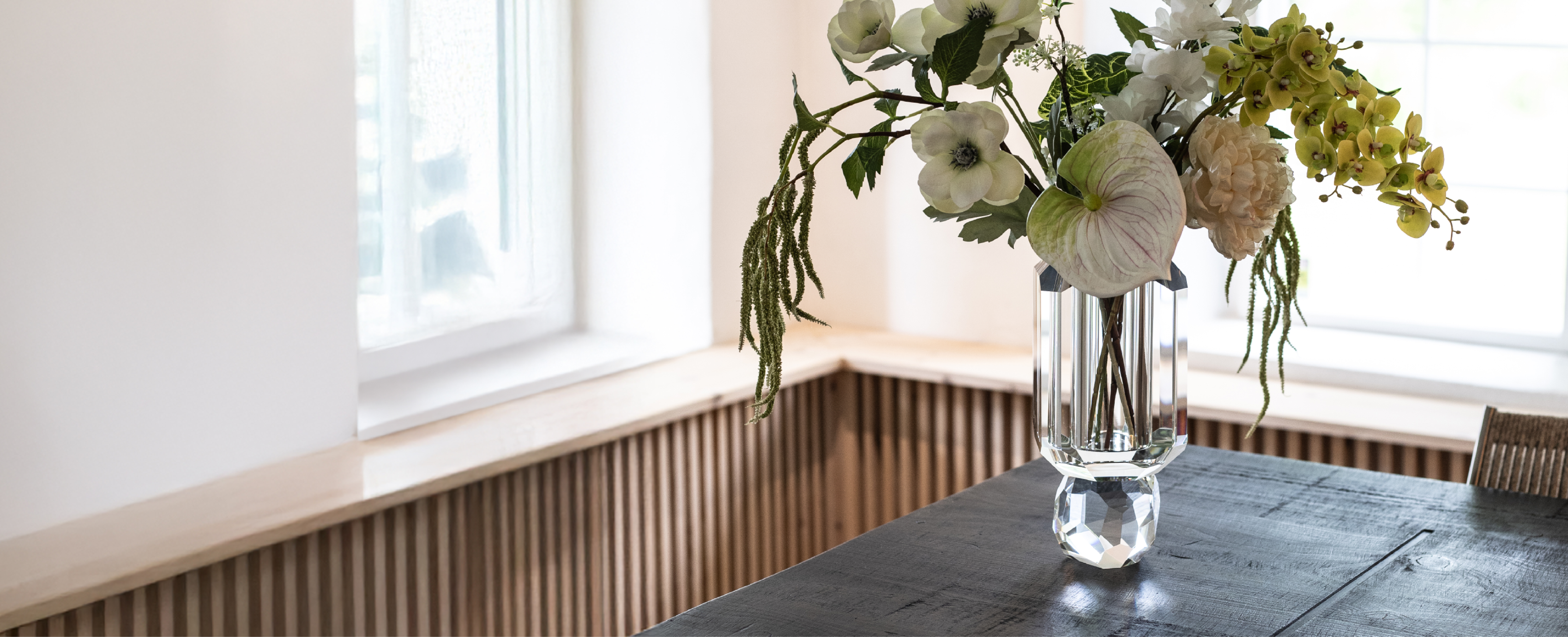 Gennemsigtig krystal vase, der står på et mørkt træ bord. I vasen er en buket kunstige blomster i lyse nuancer.