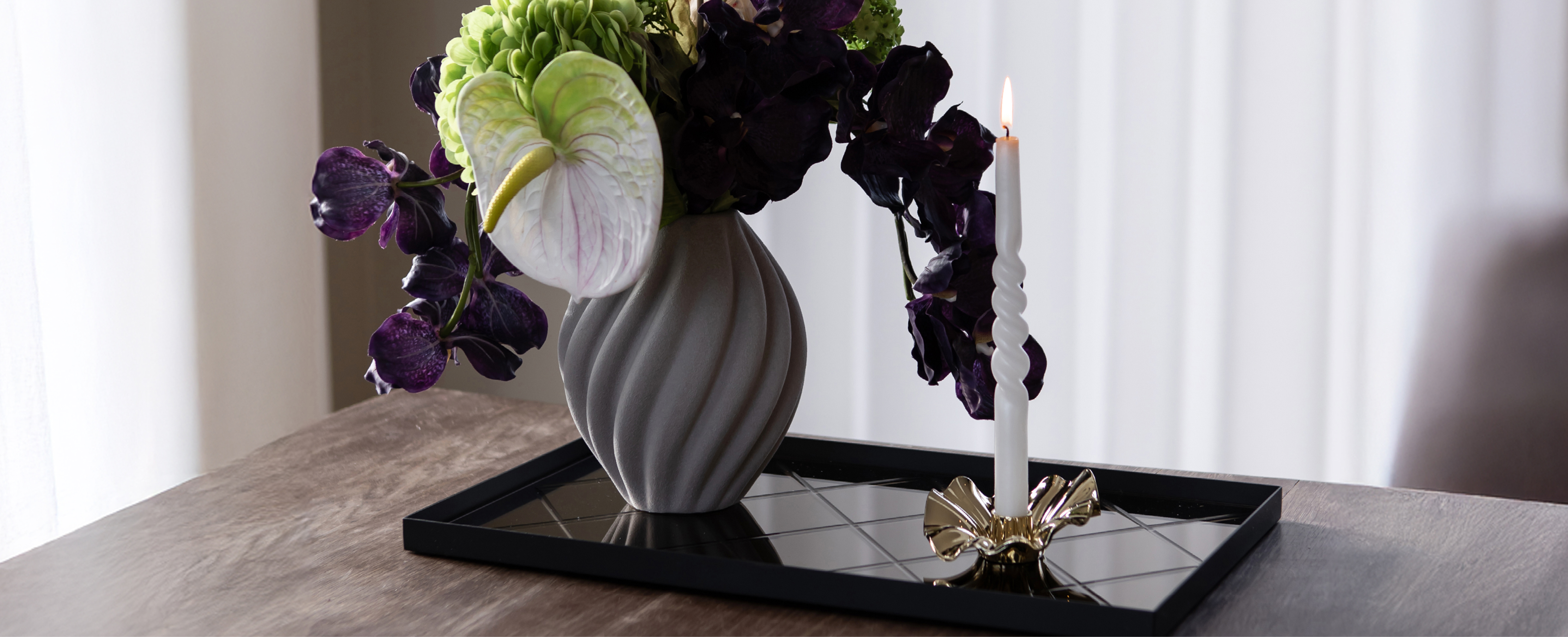Grå keramikvase med kunstige blomster, der står på en spejl bakke. På bakken står også en guld metal lysestage med hvidt snoede lys. Det hele står oven på et træ spisebord. Baggrunden er hvide gardiner.