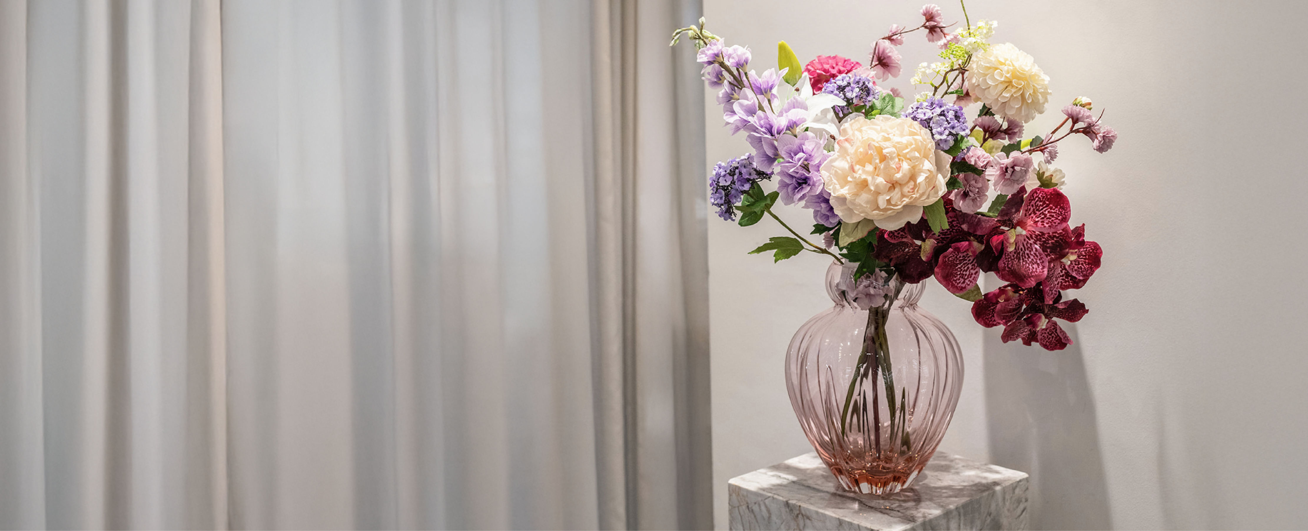 kunstig buket i rose, lilla og creme farver, der står i en lyserød glas vase på en marmor piedestal. Baggrunden er en hvid væg og grå gardiner.