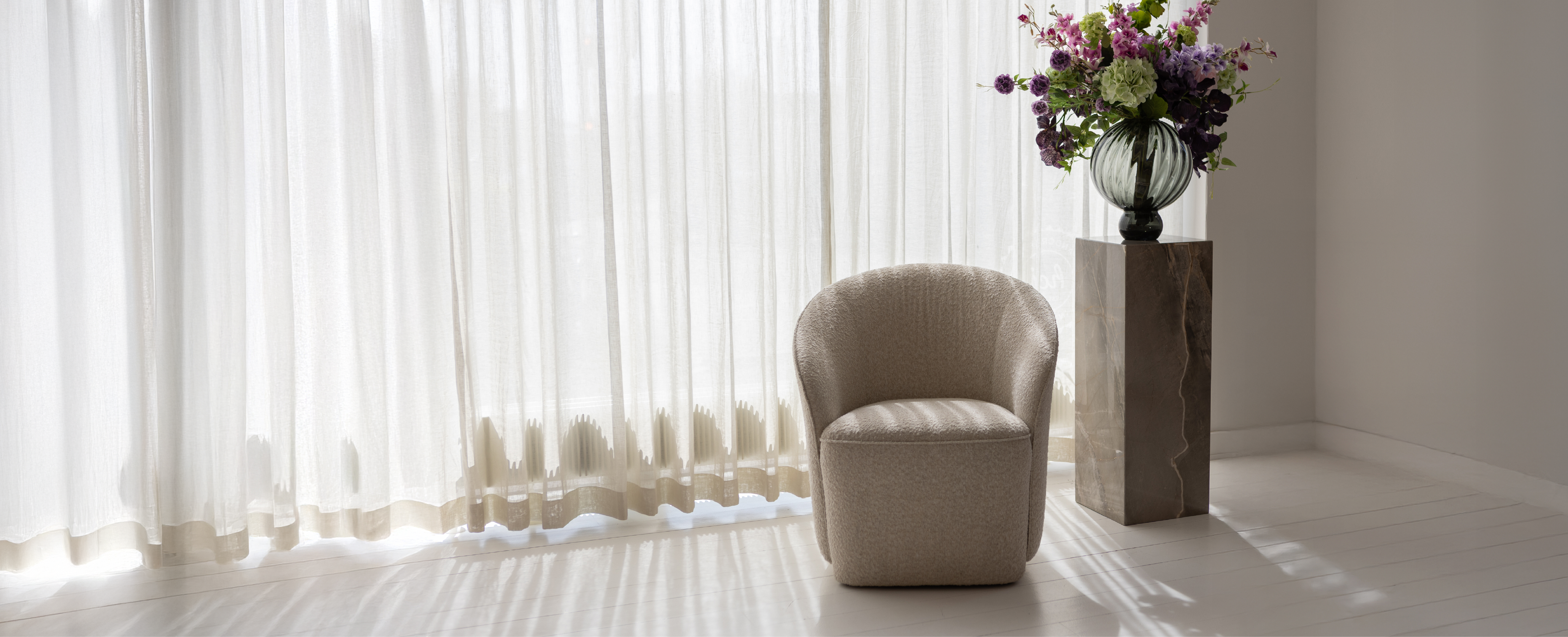 Marmor piedestal med en glas vase og kunstige blomster. Ved siden af en hvid loungestol. Baggrunden er hvid med gardiner.