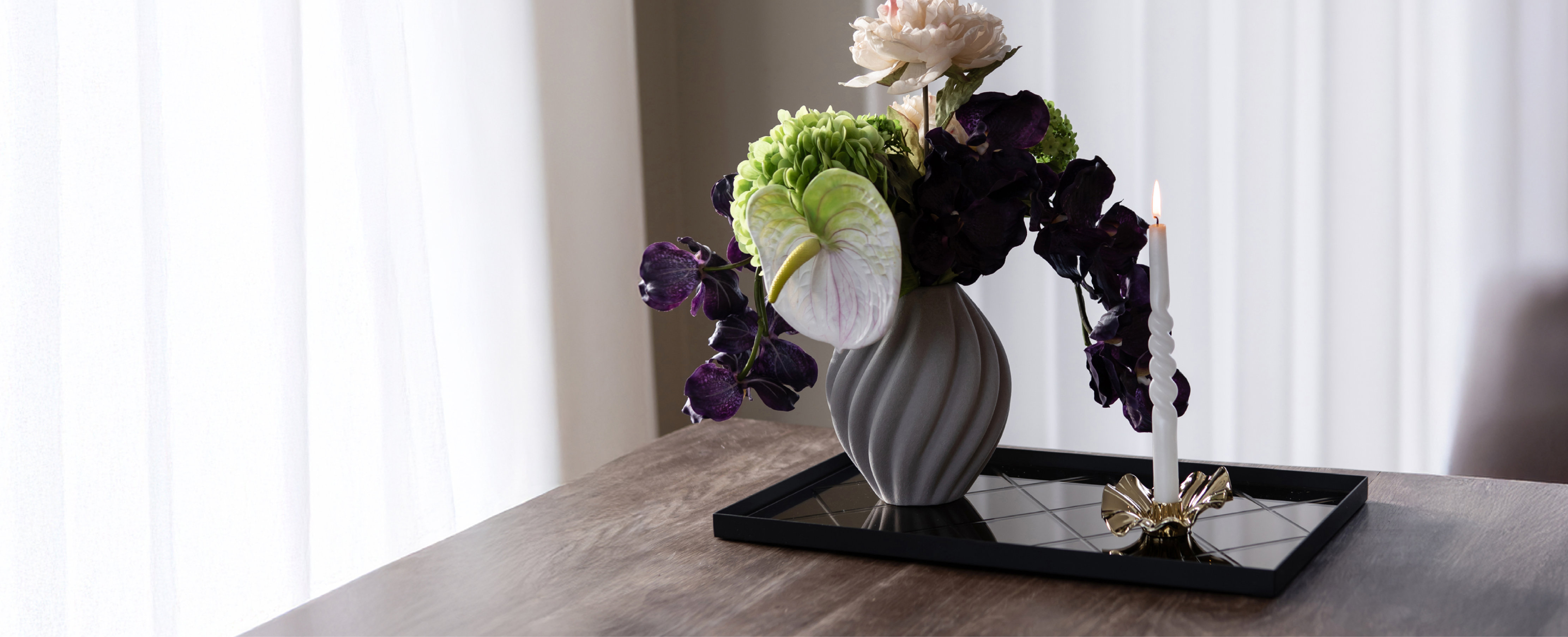 Sort spejlbakke, der står på et træ spisebord. Ovenpå bakken står en grå keramik vase med kunstige blomster og en metal lysestage i guld med et hvidt snoede lys. Baggrunden er hvide gardiner.
